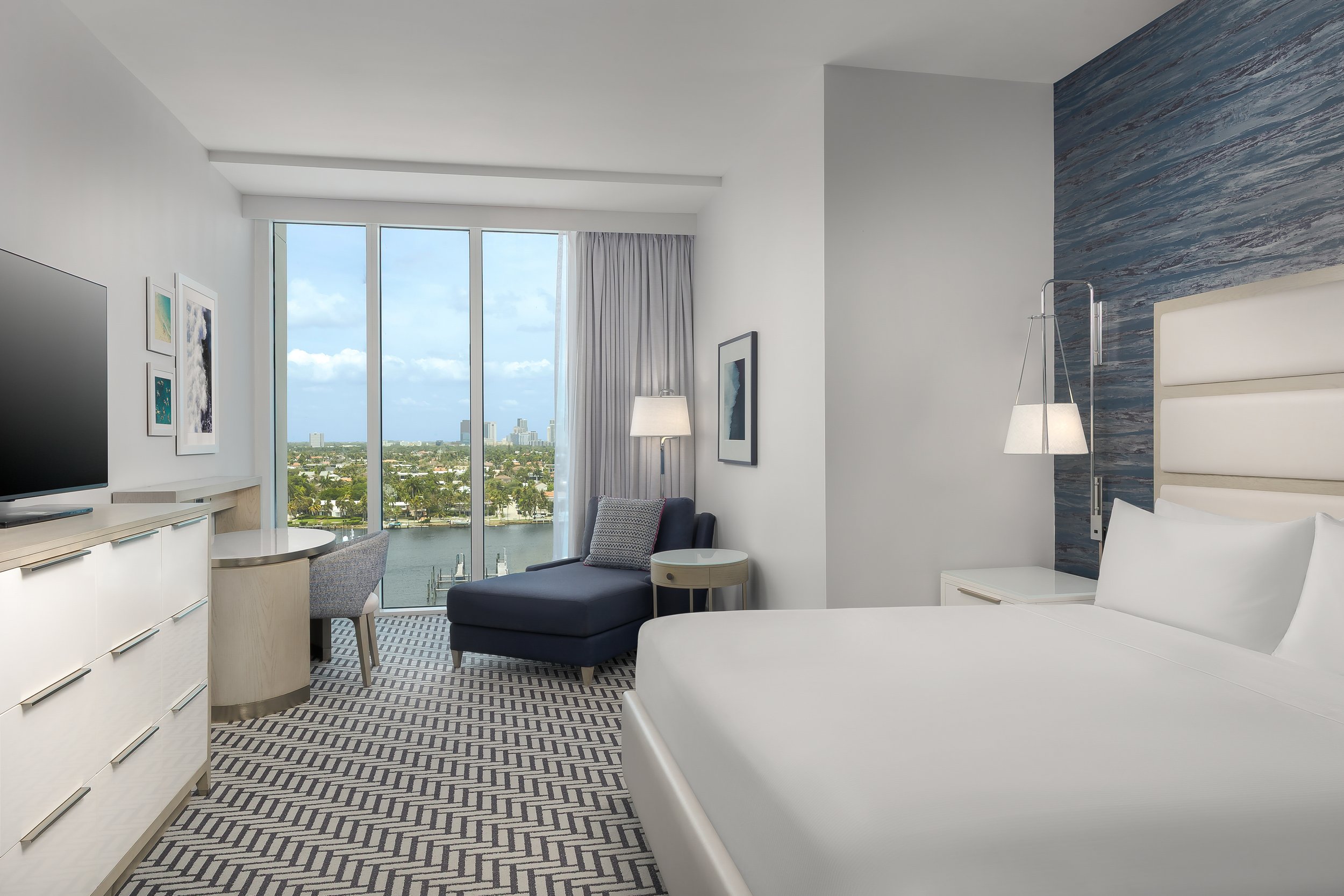 A modern hotel room with large windows offering a view of a city skyline and water. The room has a white and gray color scheme, with artwork on the walls, a television, a gray armchair with a side table, a white bed, and a floor lamp.