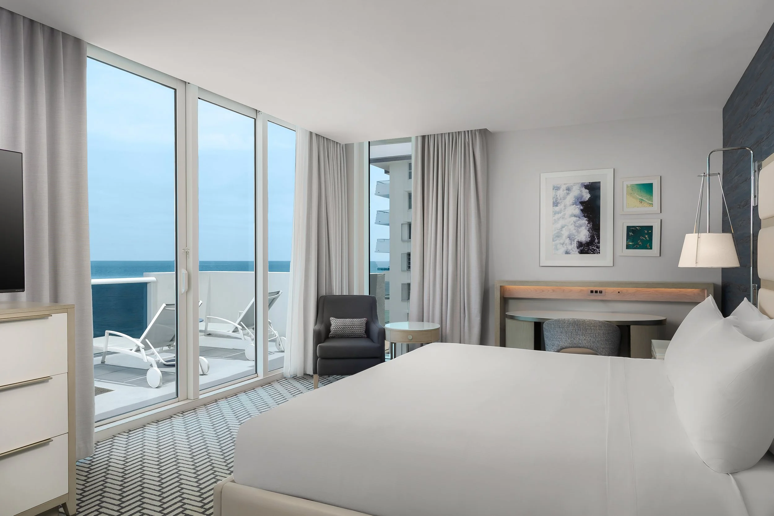 Modern hotel room with large glass sliding doors opening to a balcony overlooking the ocean. The room has white walls, artwork, a reading chair, and a bed with white linens. The balcony has lounge chairs and a view of blue sky and sea.
