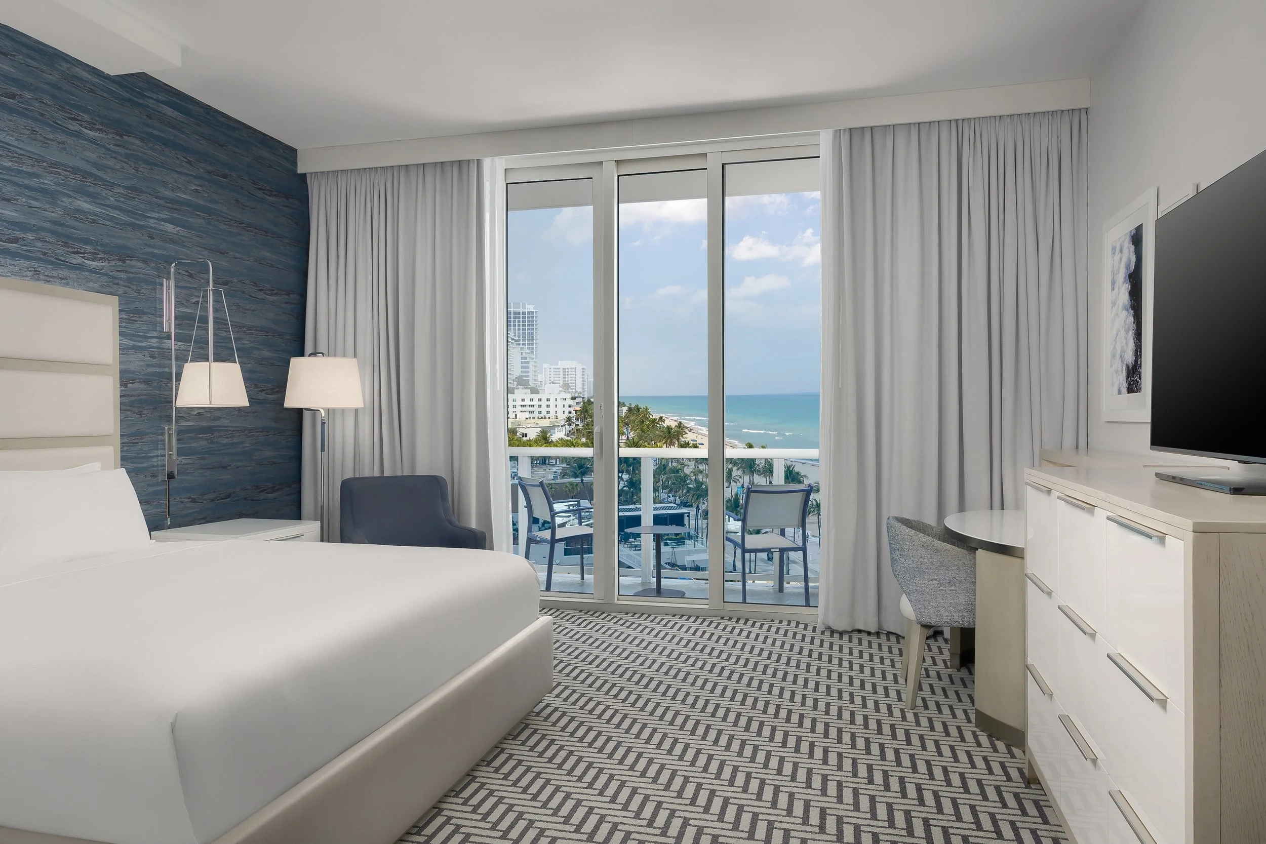 Hotel room with a large sliding glass door view of a city and ocean, white bed, blue accent wall, lamps, a small desk with a chair, and a TV.