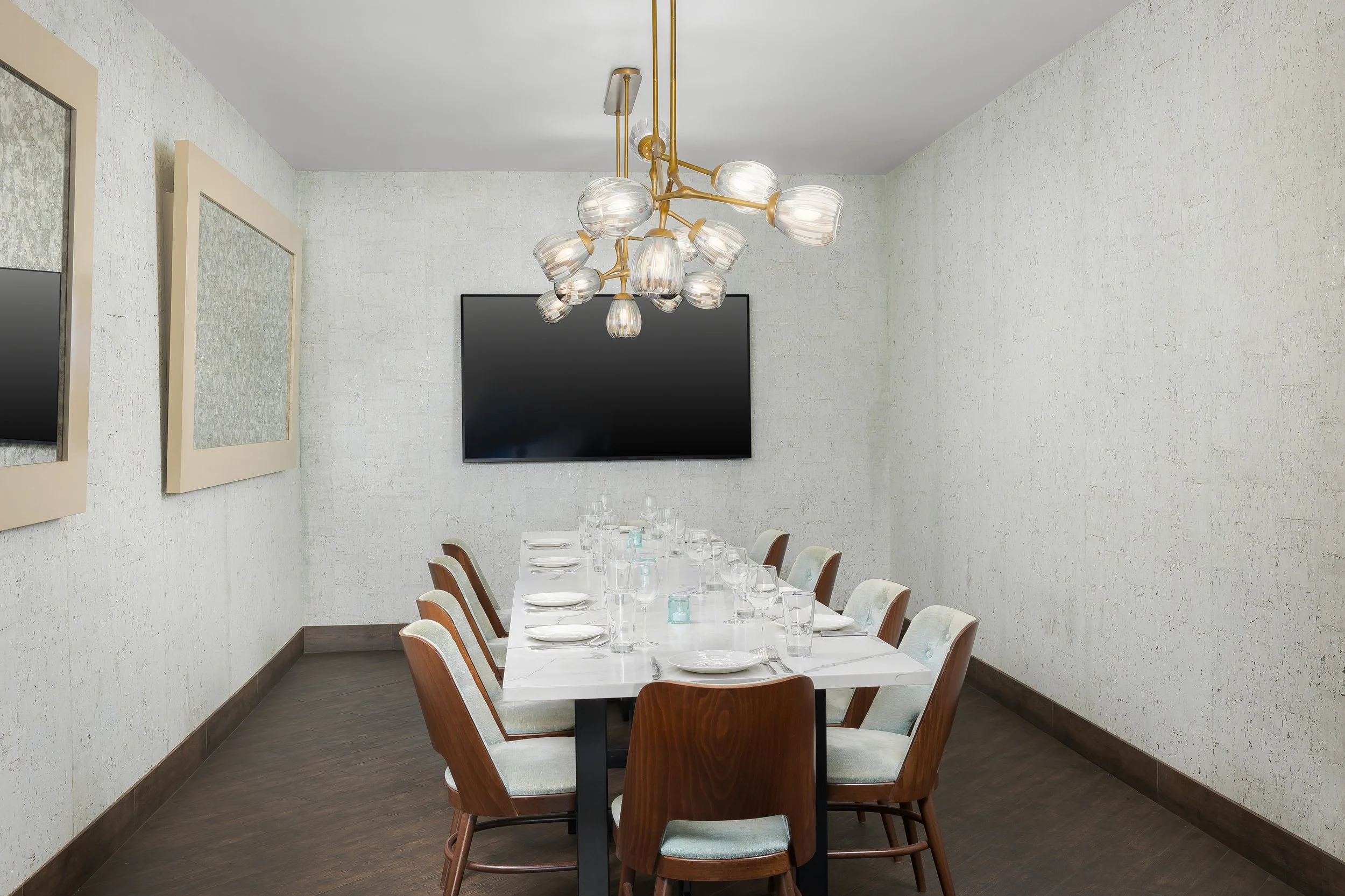 A dining room with a long white table set with plates, glasses, and napkins. Wooden chairs with light-colored cushions surround the table. A modern chandelier with multiple glass shades and gold accents hangs above the table. Two framed abstract art pieces are on the white textured walls, and a large flat-screen TV is mounted on the wall.