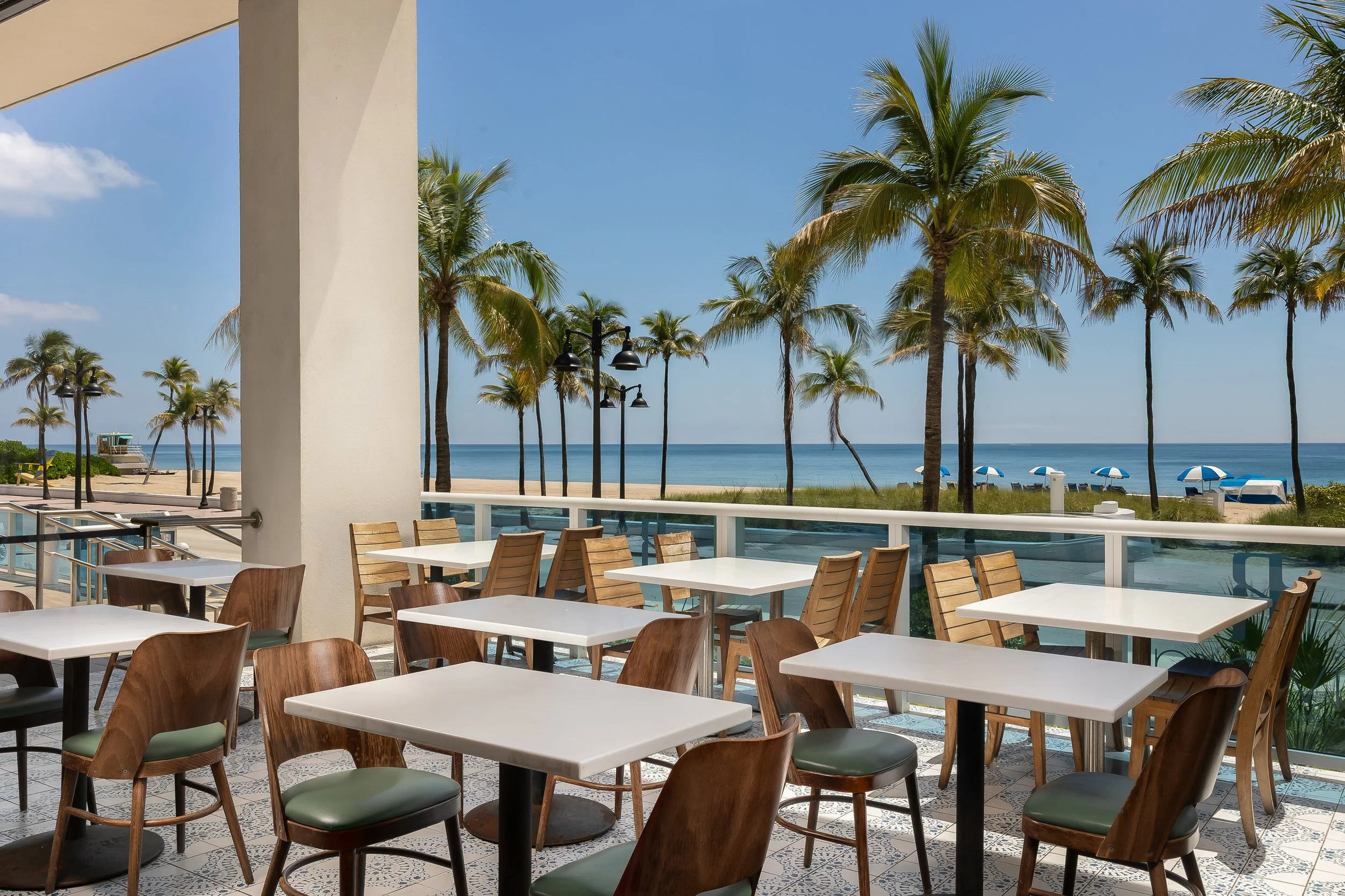 Indoor dining area with white tables and wooden chairs overlooking a beach with palm trees, blue umbrellas, and ocean in the background.