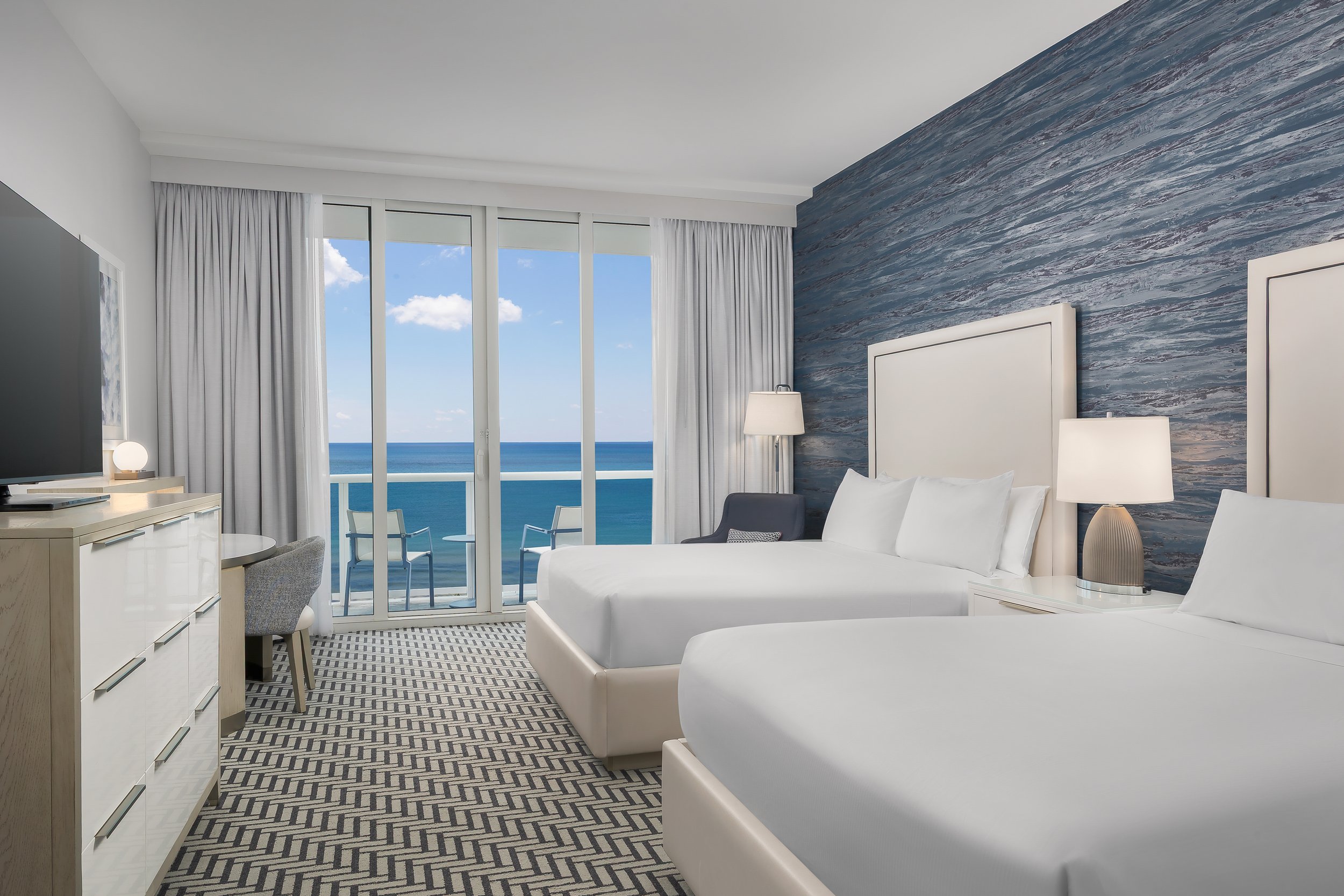 A modern hotel room with a large window overlooking the ocean and sky, featuring white and blue decor, a white bed, a dresser with a TV, a nightstand with a lamp, and a seating area.