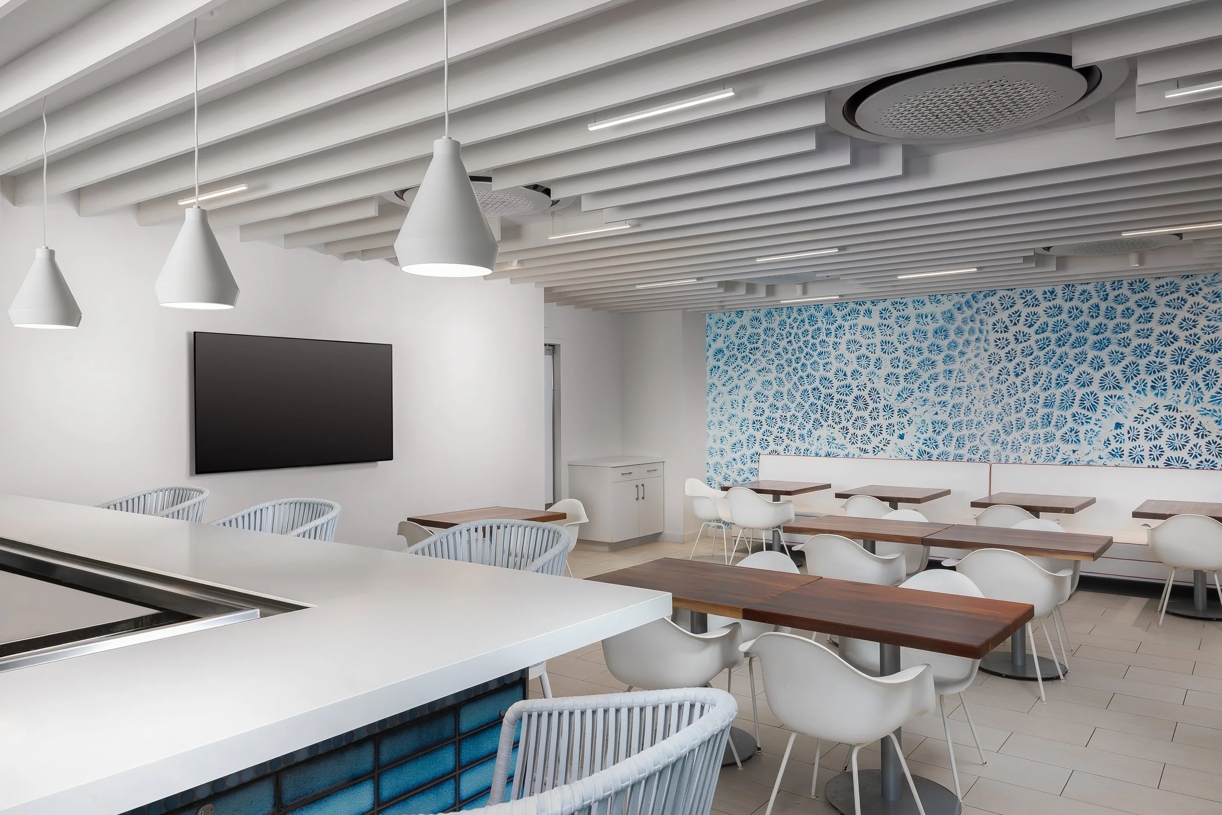 A modern, minimalist dining area with white walls and ceiling, featuring pendant lights, a wall-mounted TV, and a blue and white patterned accent wall. There are wooden tables and white chairs arranged around the space.