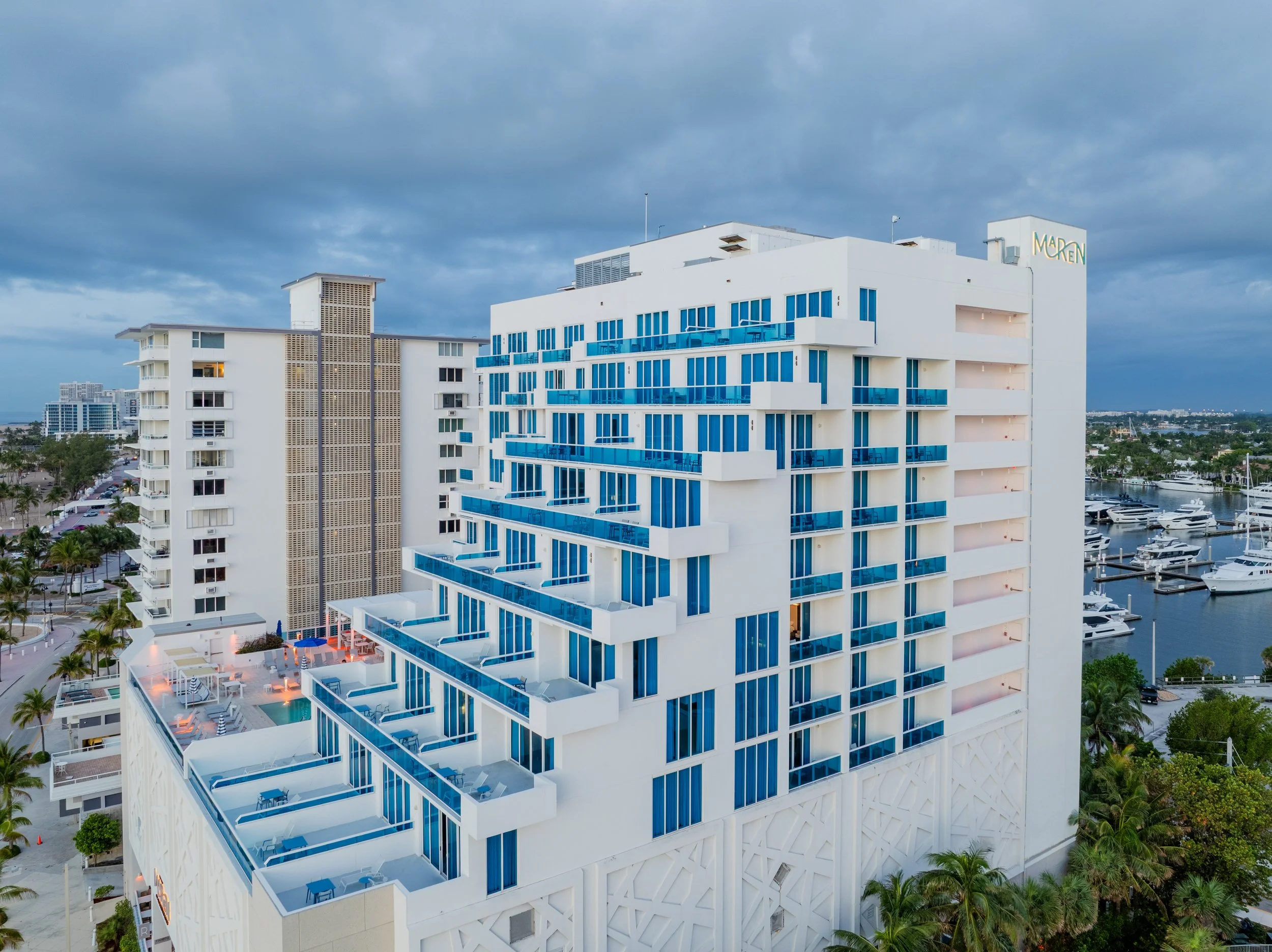 An upscale white modern hotel building with blue-tinted glass balconies and a rooftop pool area, overlooking palm trees, a marina with yachts, and a cloudy sky.