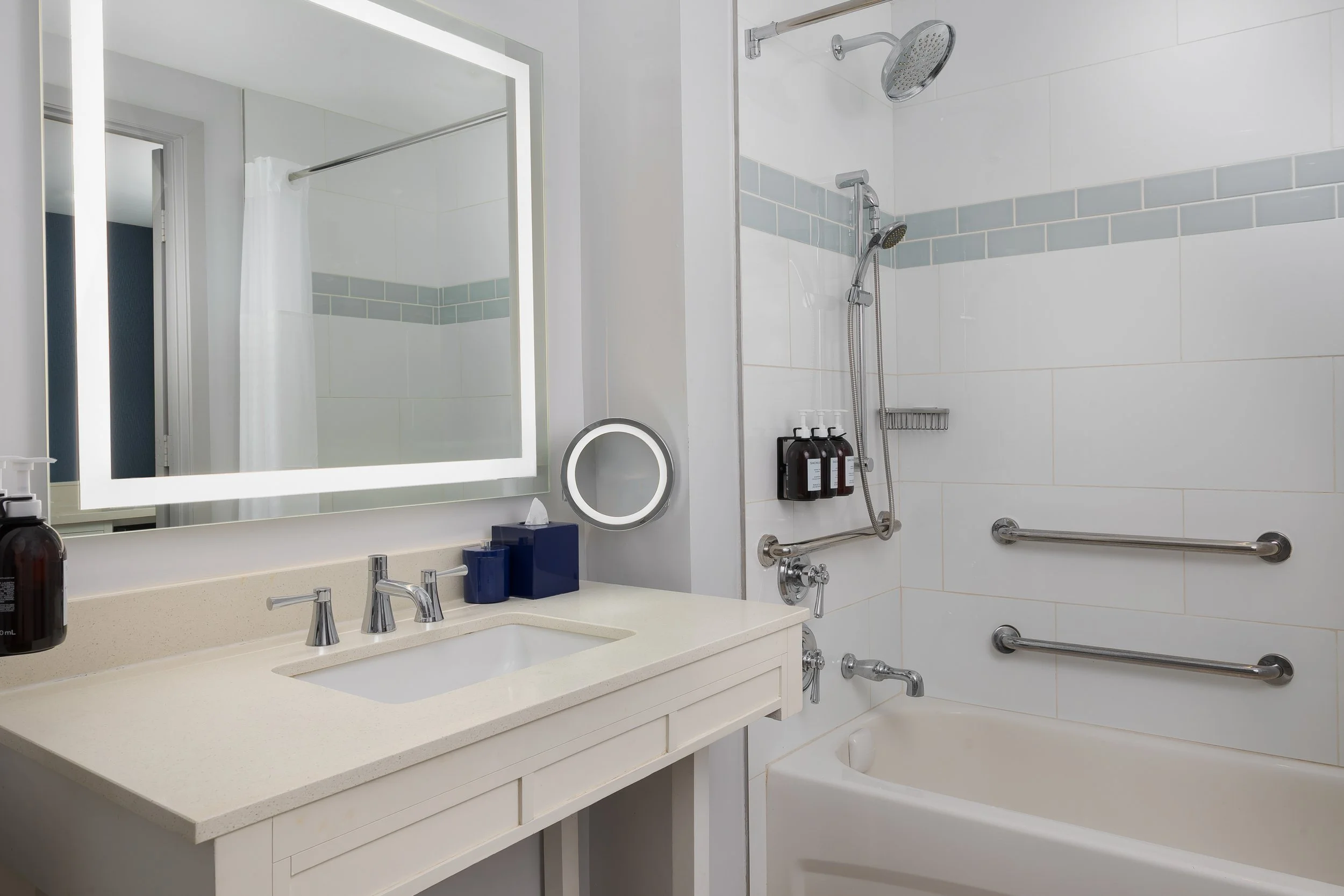 A bathroom with a white vanity, large mirror, soap dispenser, and hand towels. Shower area has a mounted rainfall showerhead, a handheld showerhead, and three black bottles on a wall-mounted shelf. The bathtub has safety grab bars installed.