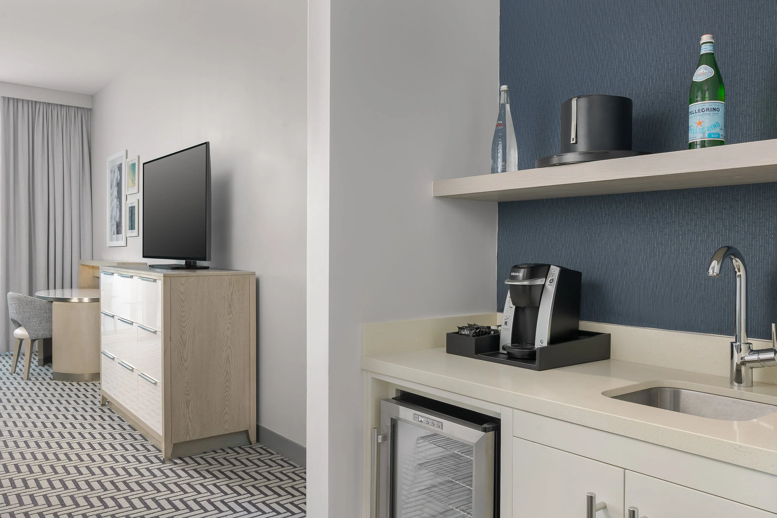 A kitchen corner with a white countertop, coffee maker, small refrigerator, and a blue textured wall shelf with water and sparkling water bottles, in a modern living space.