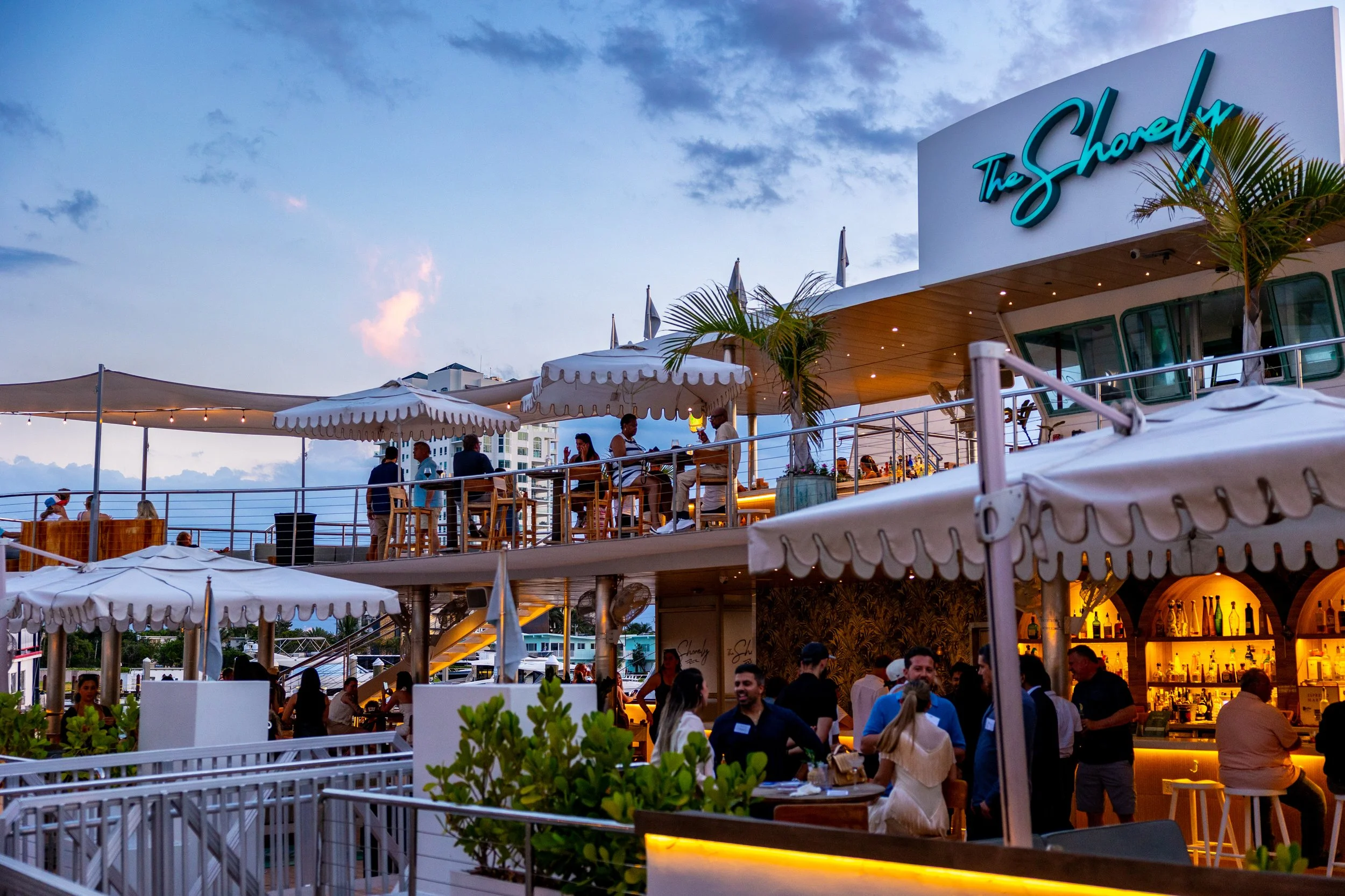 A rooftop bar and restaurant named The Shorely, with people dining and socializing under umbrellas during the evening. The sky is partly cloudy with the sun setting, and there are palm trees and a city skyline in the background.