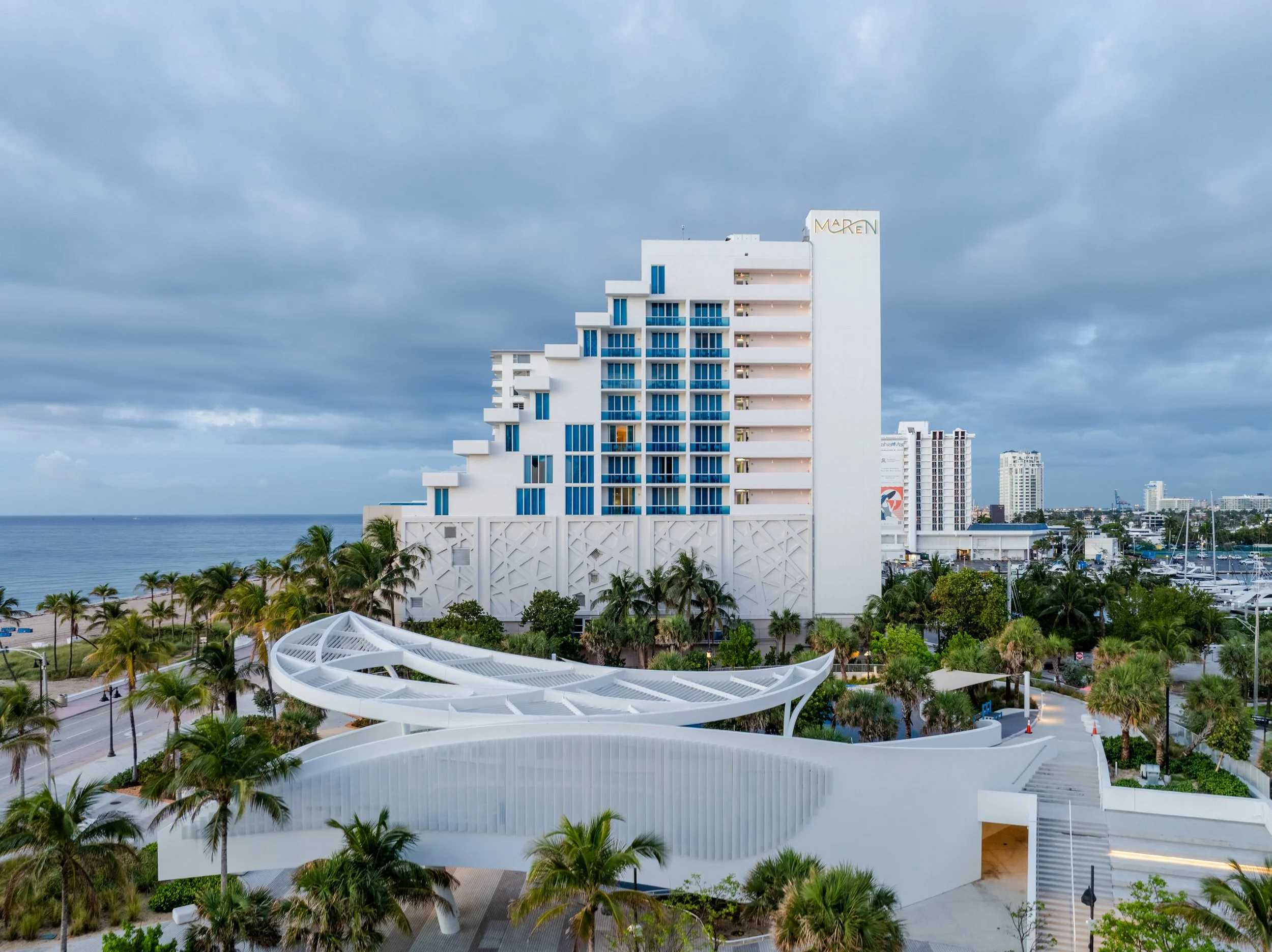 A modern white hotel building with dark blue windows and balconies, situated near the coast with palm trees in the foreground and a cloudy sky overhead.