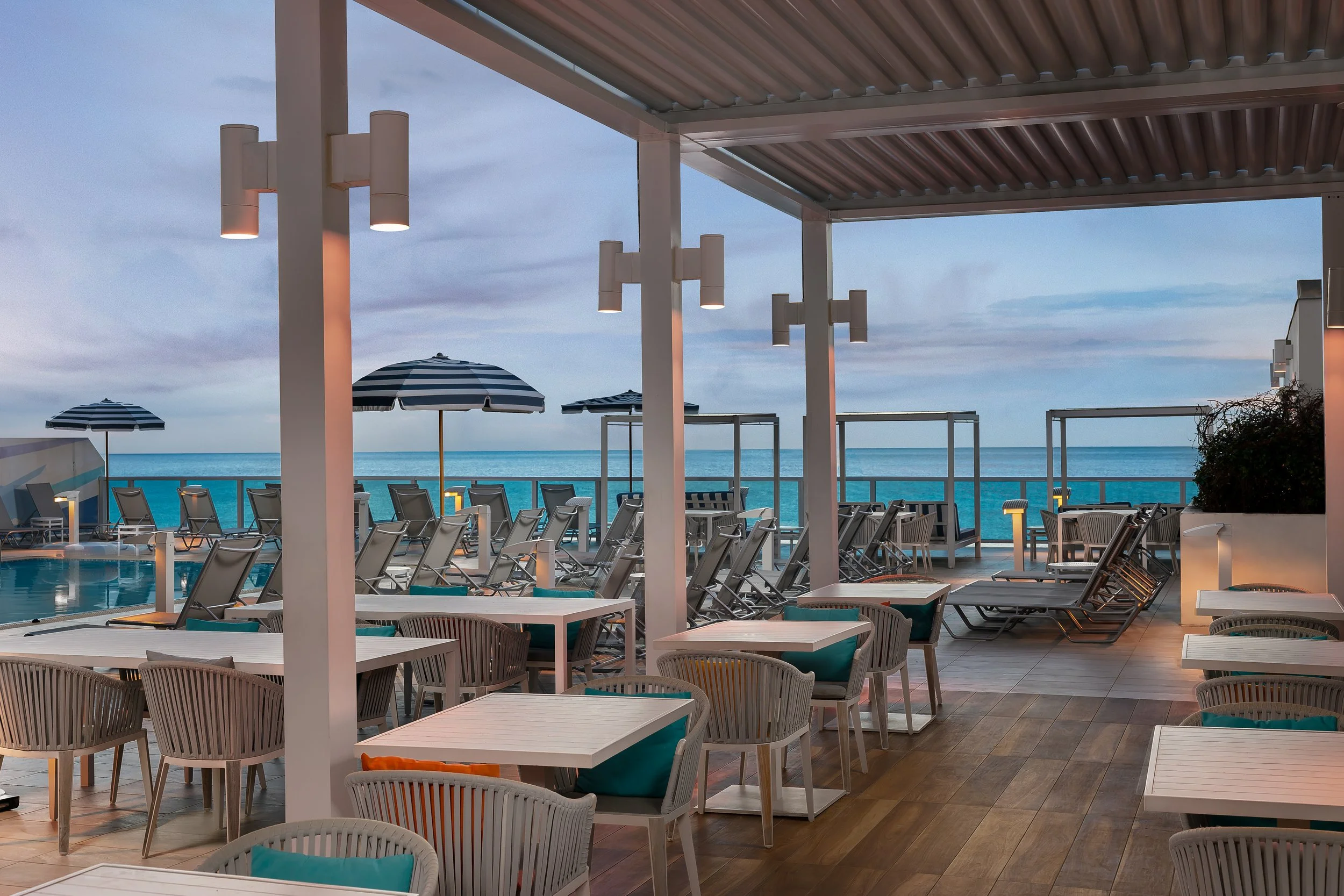 An outdoor poolside area at a hotel or resort with chairs, tables, and umbrellas overlooking the ocean, under a covered patio with modern lighting and a view of the sky.