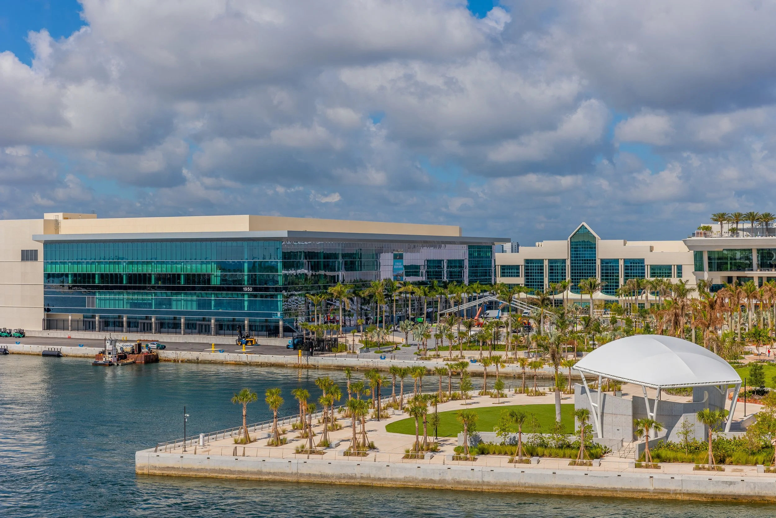 A modern building with large glass windows, a waterway in the foreground, palm trees, and a partly cloudy sky.