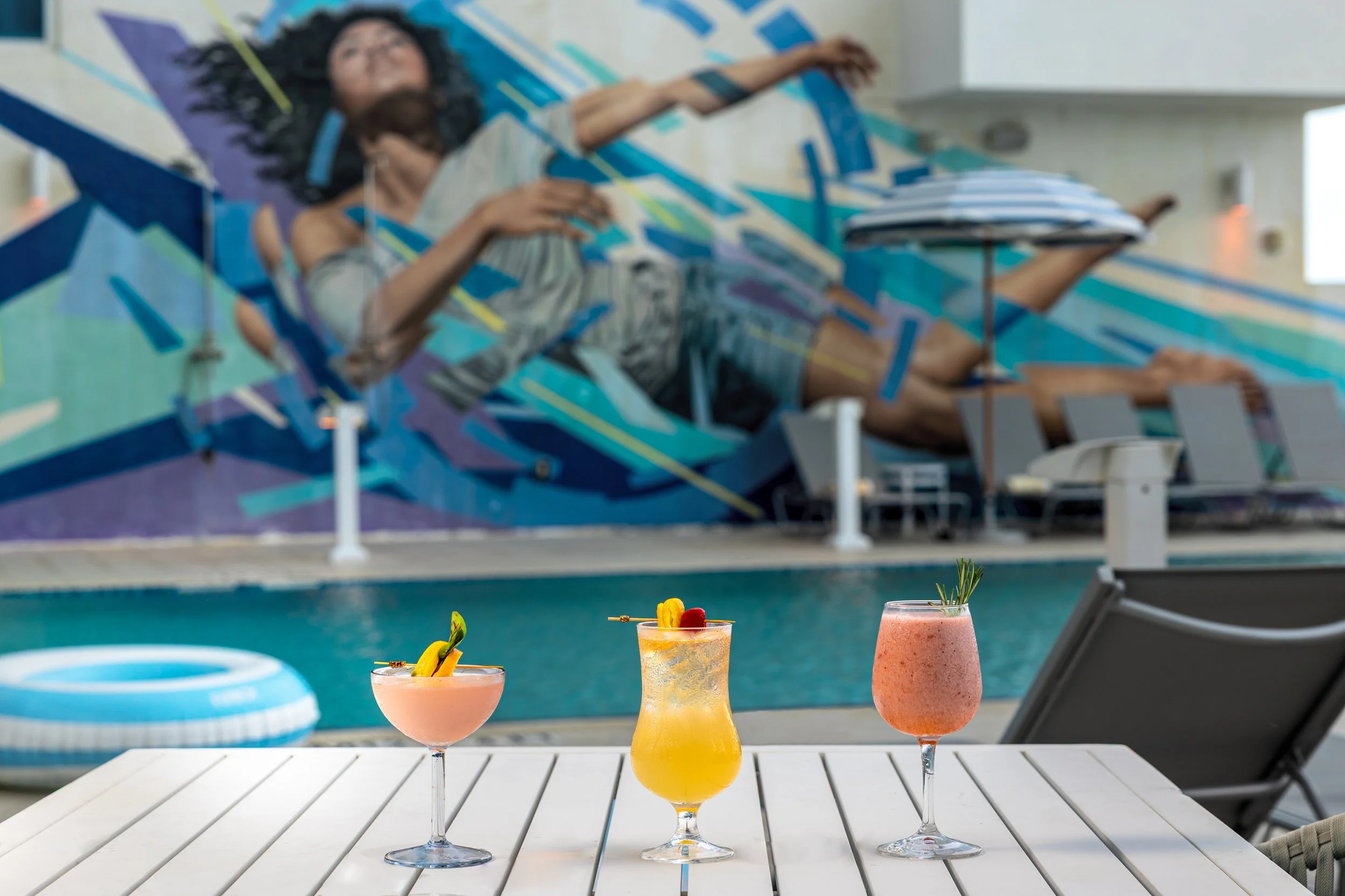 Three colorful cocktails on a white poolside table with a swimming pool and a mural with a woman in the background.