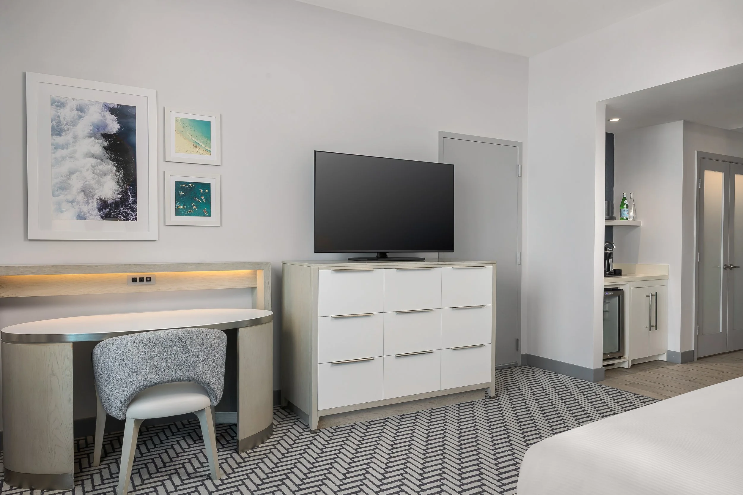 Modern hotel or apartment room with white walls, a mounted black flat-screen TV, and framed beach and ocean artwork on the wall. Features a small table with a grey chair, a white cabinet, a mini fridge, and a kitchenette area with bottles on a shelf.