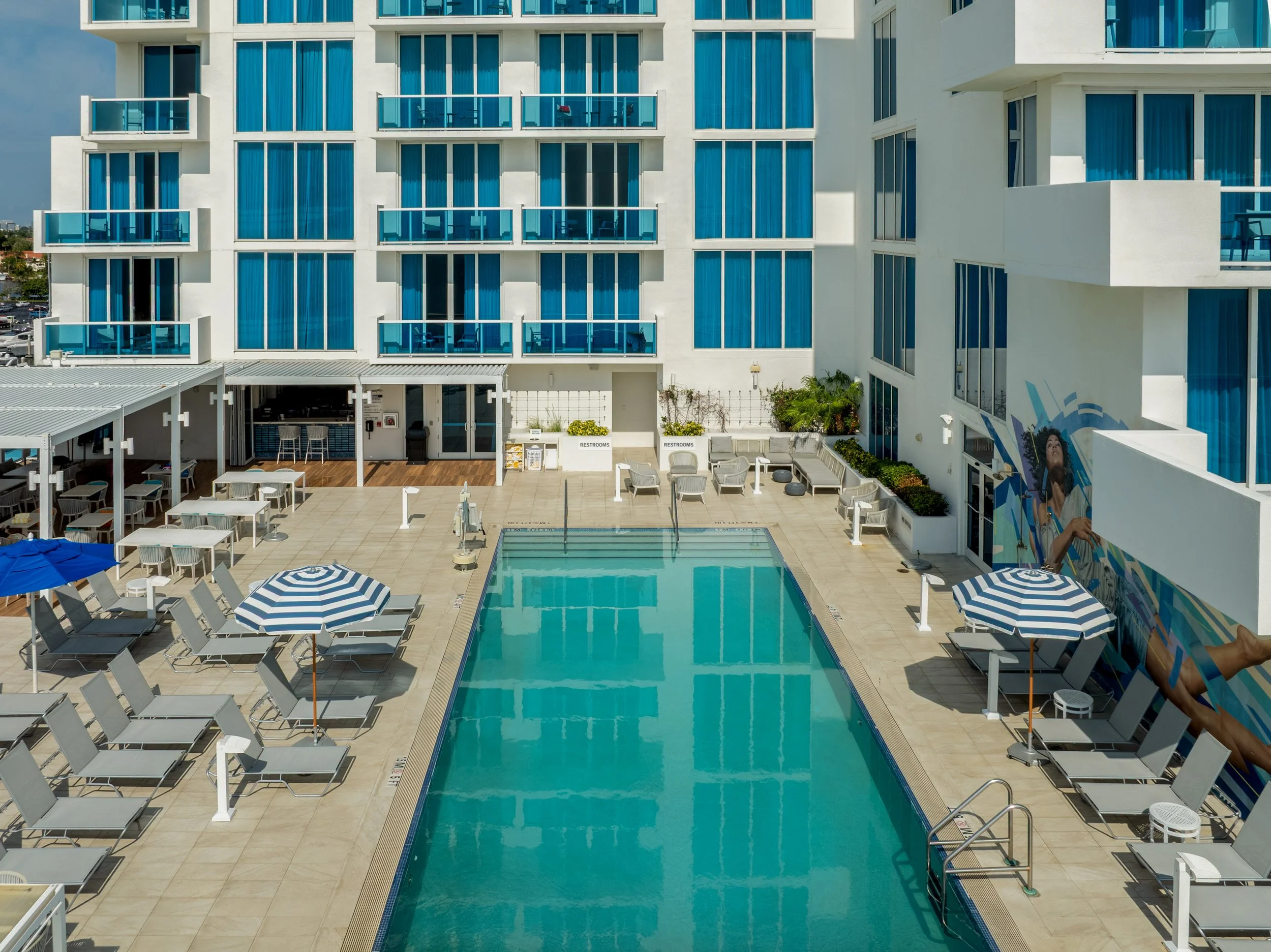 View of a swimming pool area at a hotel or resort, with lounge chairs, umbrellas, and a multi-story building with blue-tinted windows and balconies in the background.
