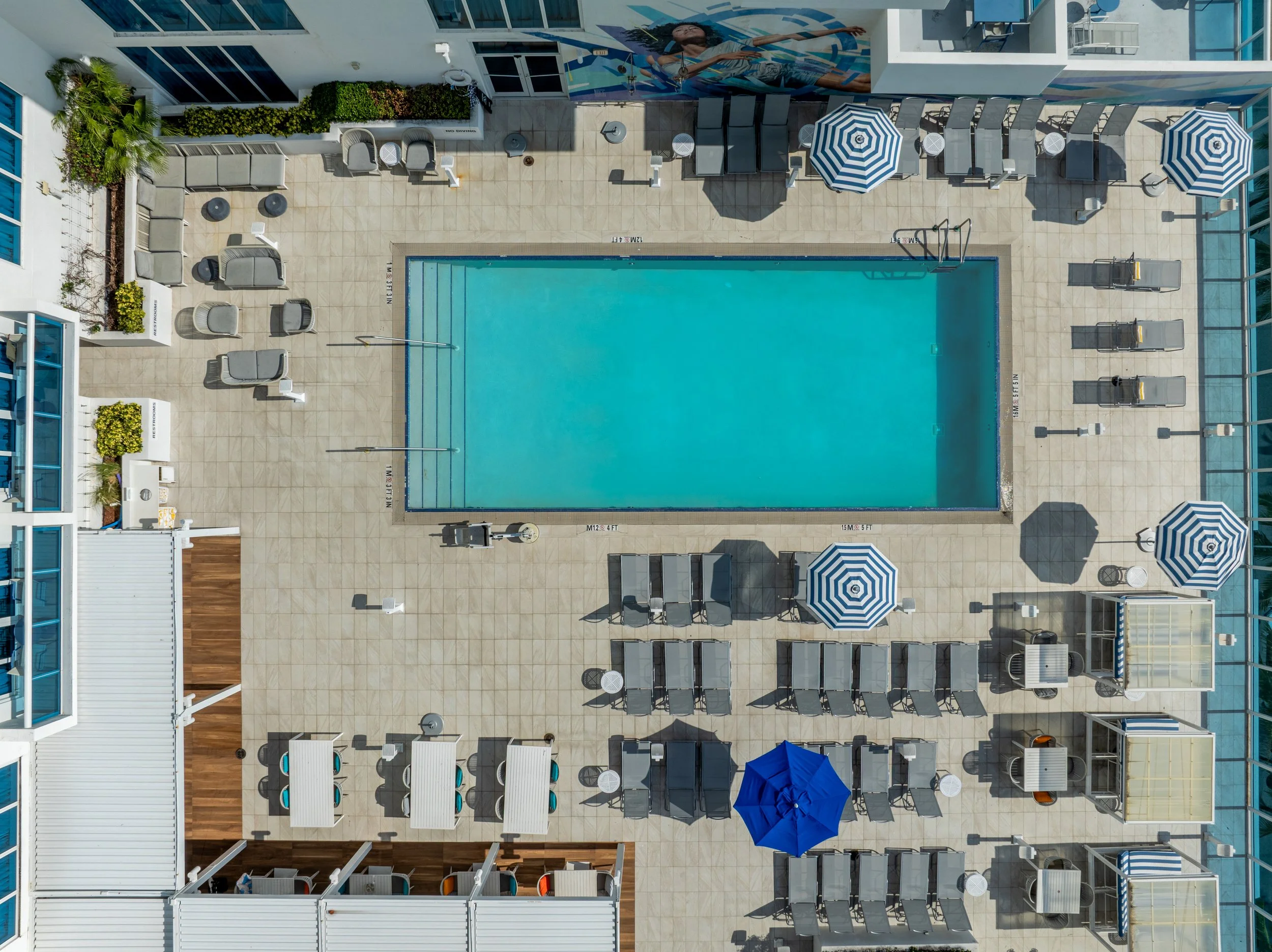 Top-down view of a swimming pool area with lounge chairs, umbrellas, and a mural of a woman swimming painted on the wall.