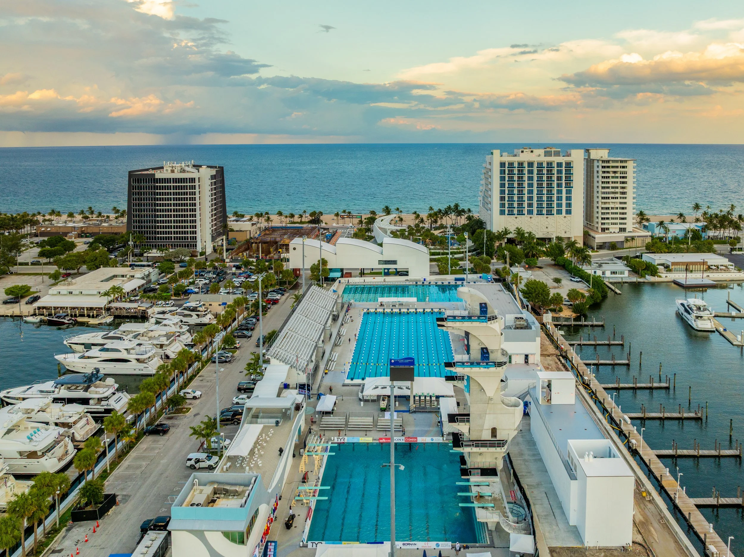View of a sports stadium with a swimming pool and tall buildings by the beach, with yachts in the water and palm trees lining the streets.