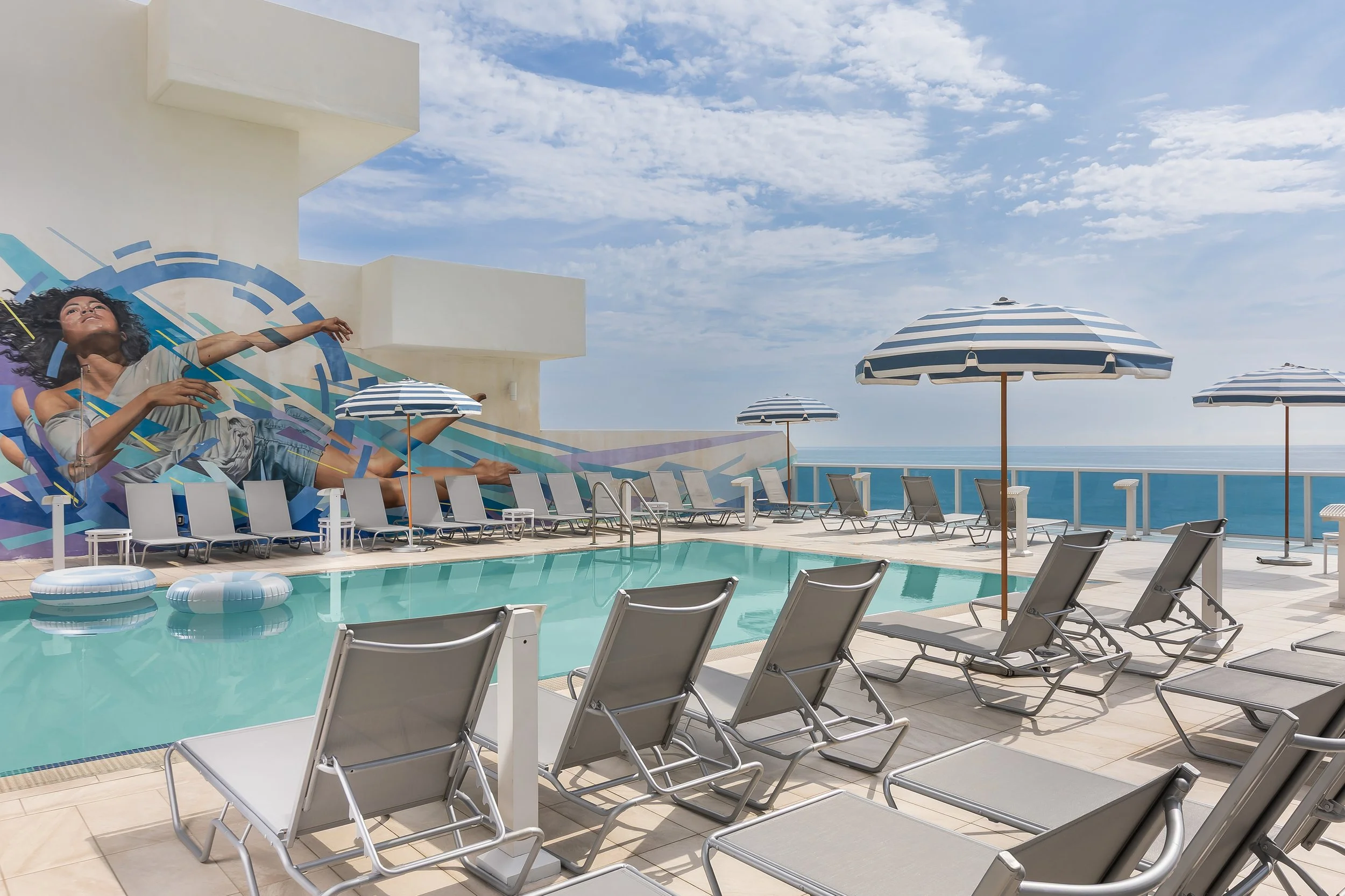 Pool area with lounge chairs, striped umbrellas, and a mural of a woman reaching out, overlooking the ocean under a partly cloudy sky.