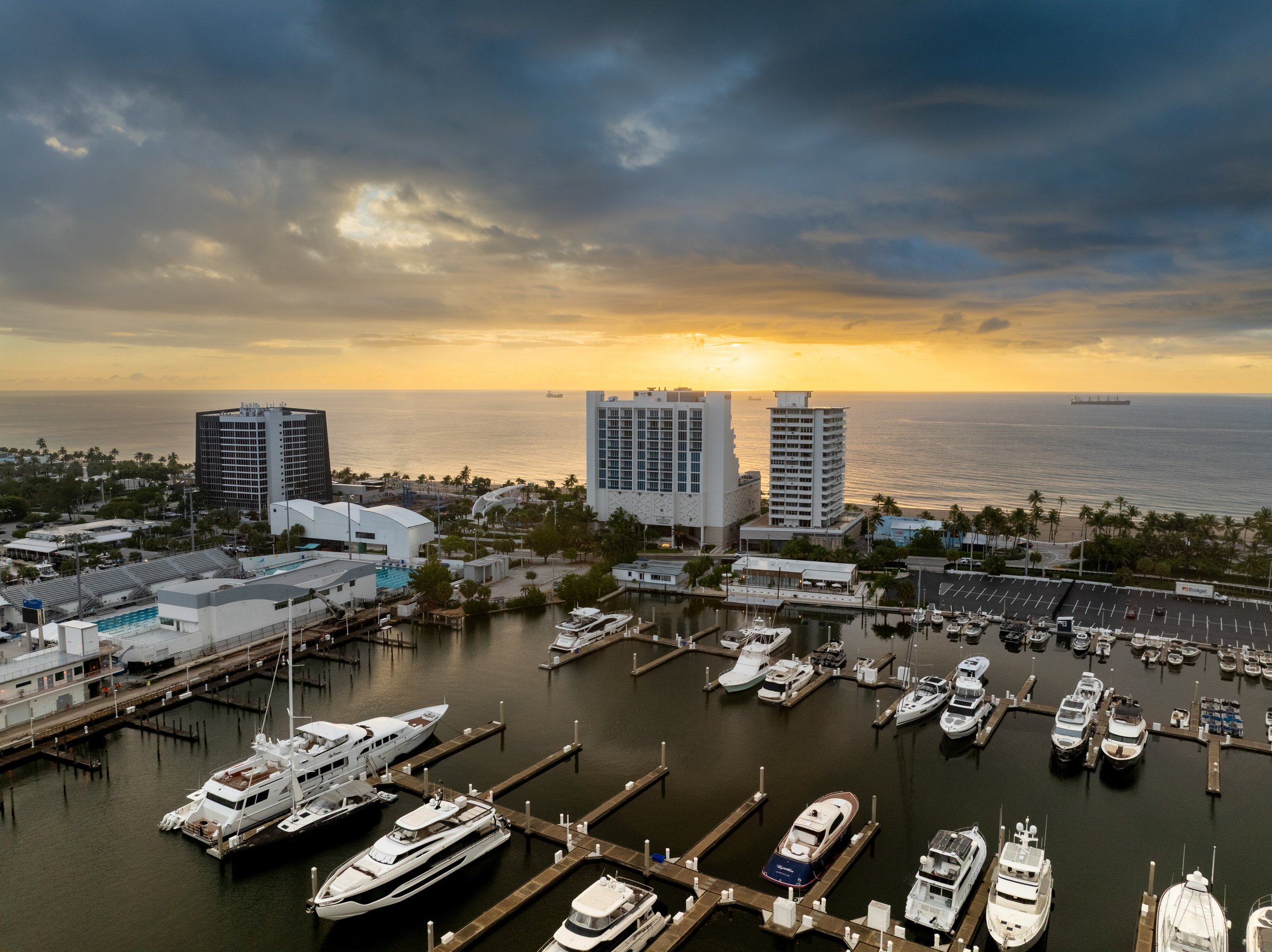 View of a marina with yachts and boats docked, high-rise buildings along the coast, and a cloudy sky with the sun setting over the ocean in the background.