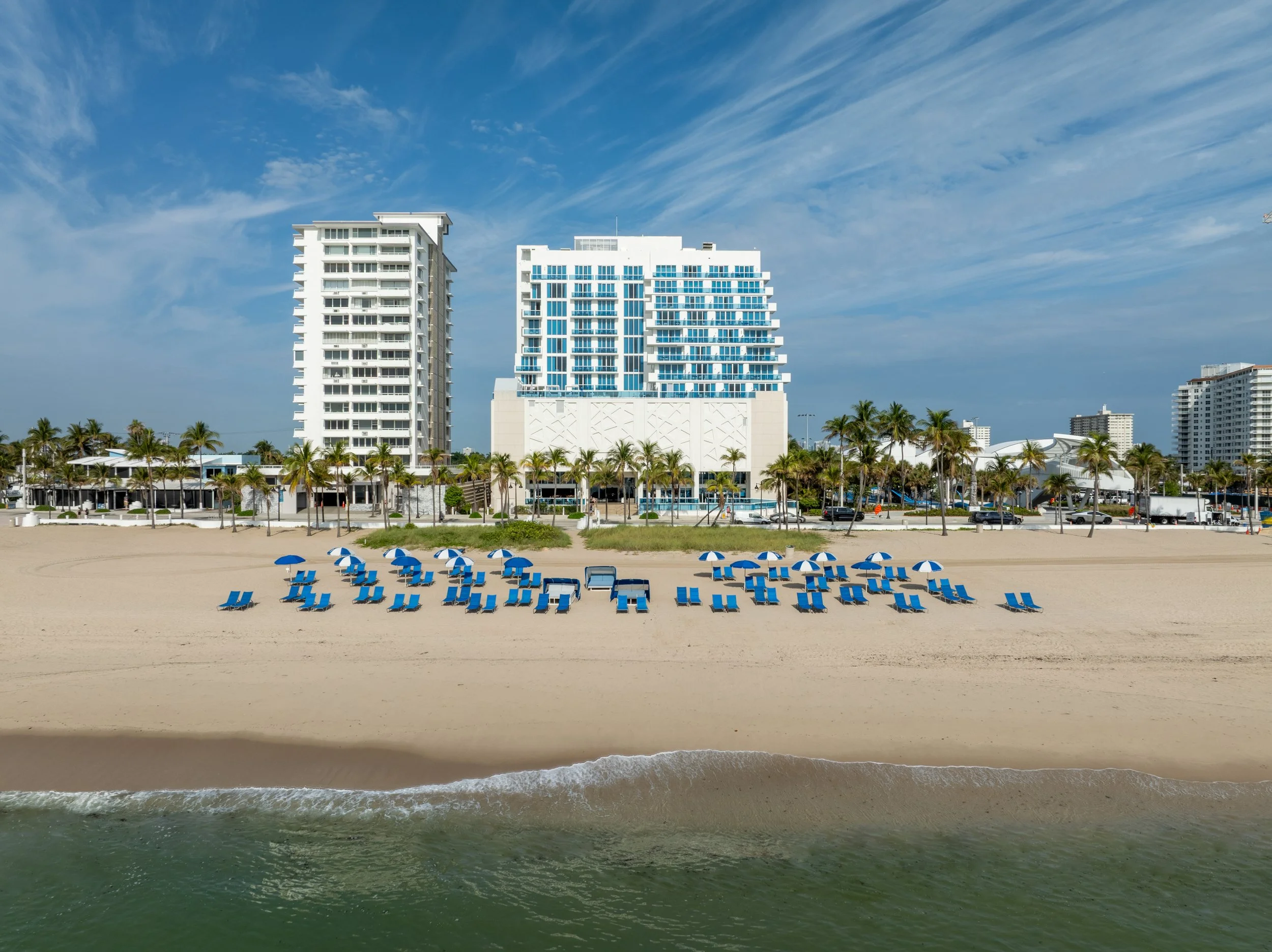 A beach with blue umbrellas and lounge chairs in front of high-rise buildings and palm trees, under a partly cloudy sky.