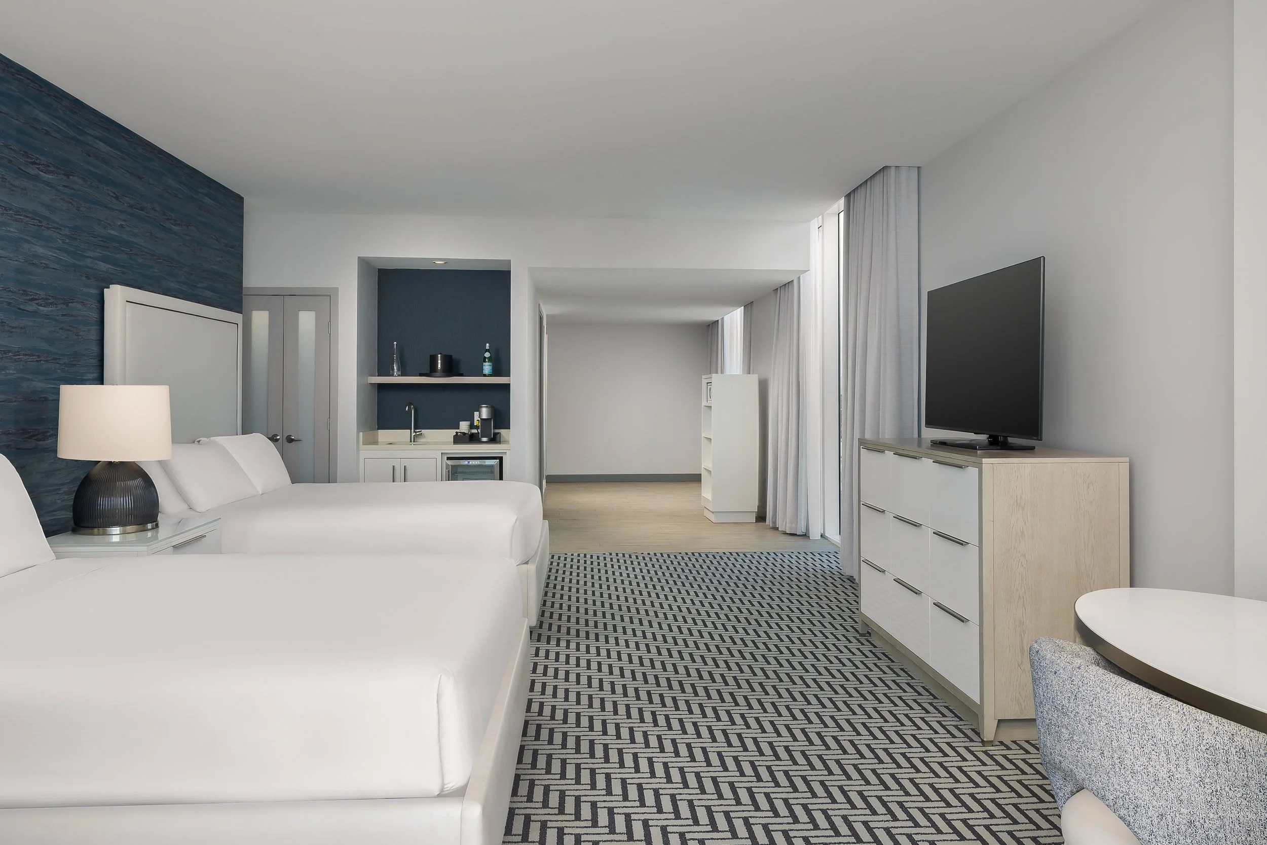 Interior of a modern hotel room featuring white sofas, a patterned rug, a dresser with a TV, and a kitchenette with blue accent walls, white cabinetry, and a small table.