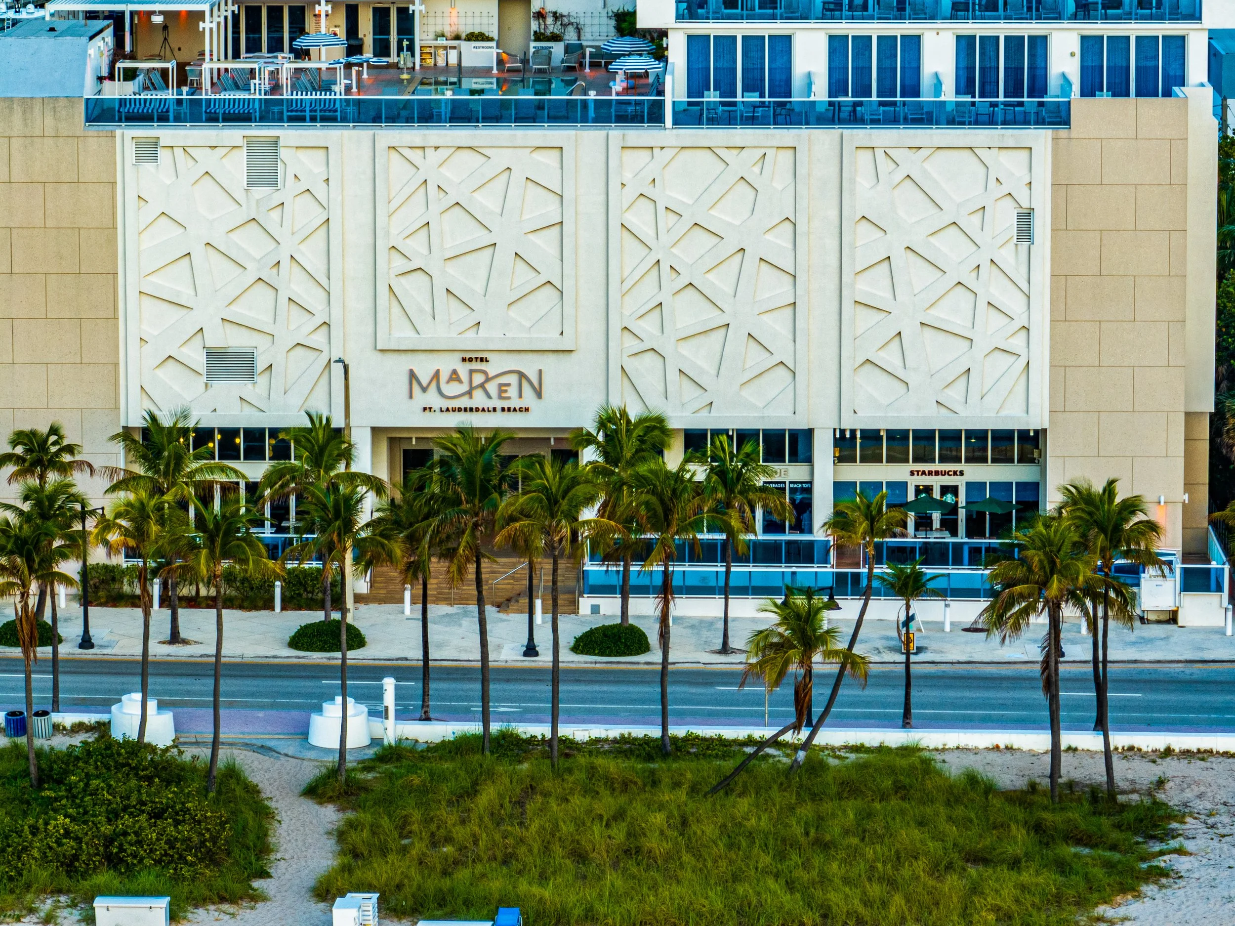 Exterior view of the Hotel Maren Ft. Lauderdale Beach with palm trees in the foreground, a paved walkway, and a road.