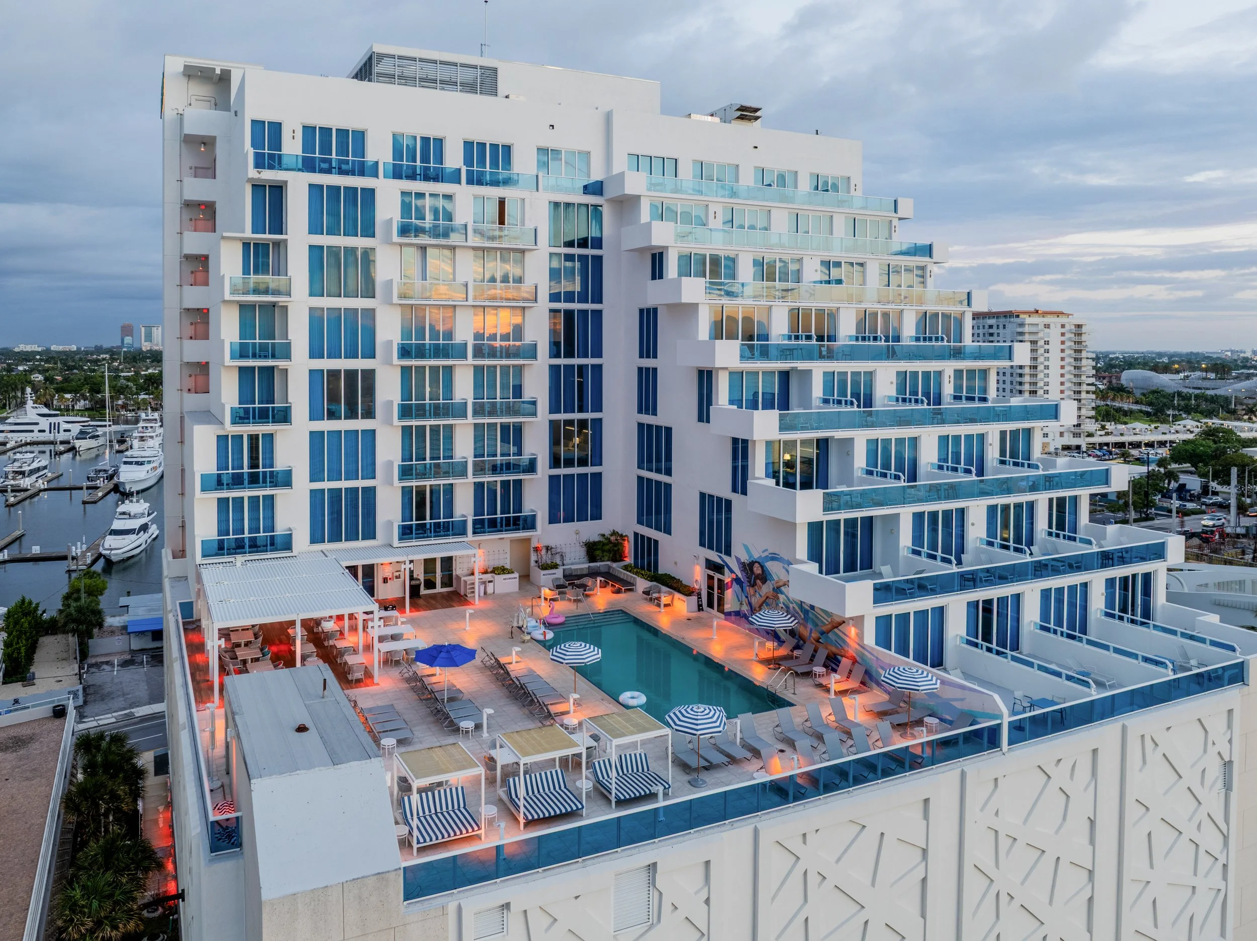 A modern white multi-story hotel building with blue-tinted glass balconies overlooking a marina with yachts and boats, and a swimming pool area at the base with lounge chairs, umbrellas, and outdoor seating. Cityscape and cloudy sky in the background.