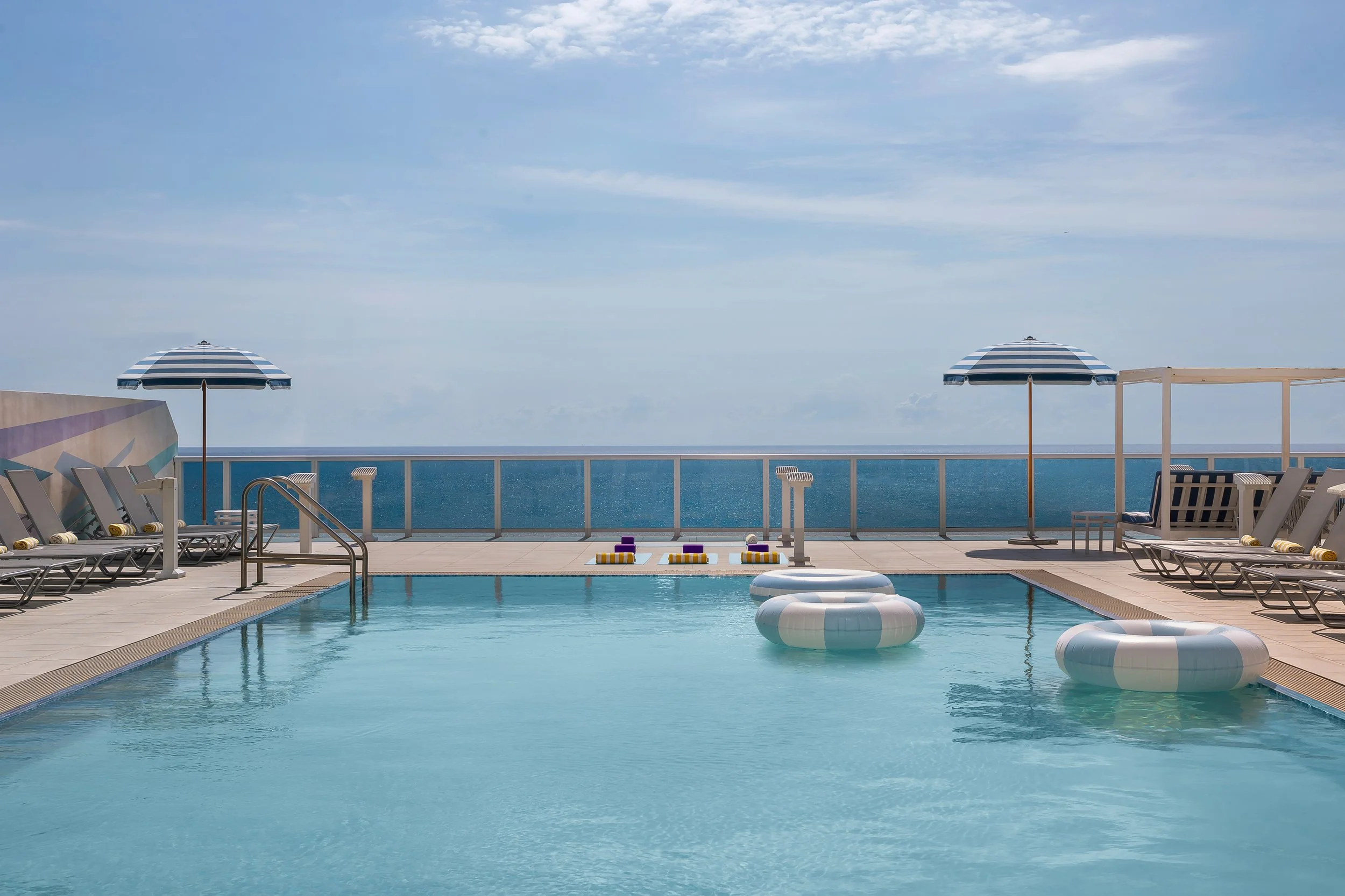 A rooftop swimming pool with pool floats, surrounded by lounge chairs and striped umbrellas, overlooking the ocean under a blue sky.