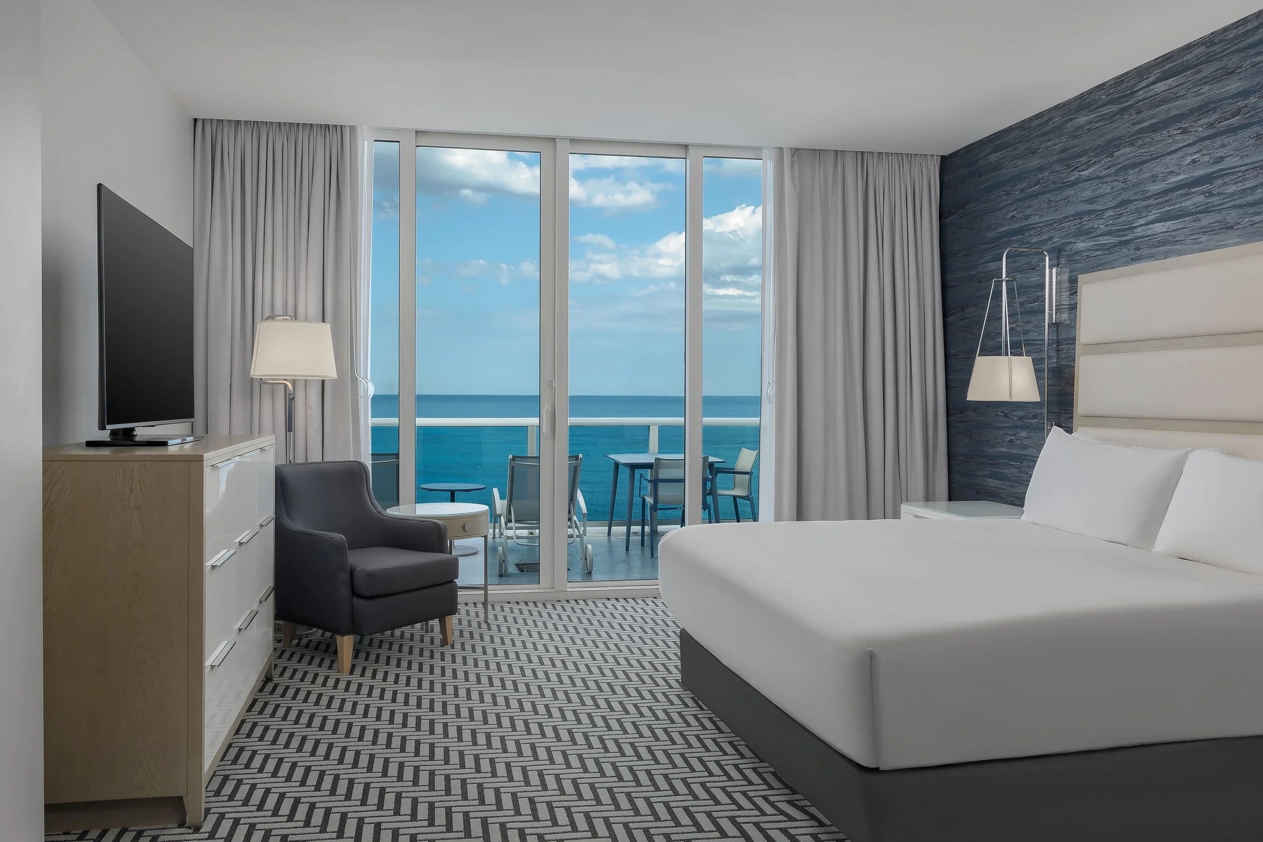 Hotel room with a large window overlooking the ocean, featuring a bed, a TV on a wooden dresser, a gray armchair, a nightstand, and a balcony with outdoor furniture.