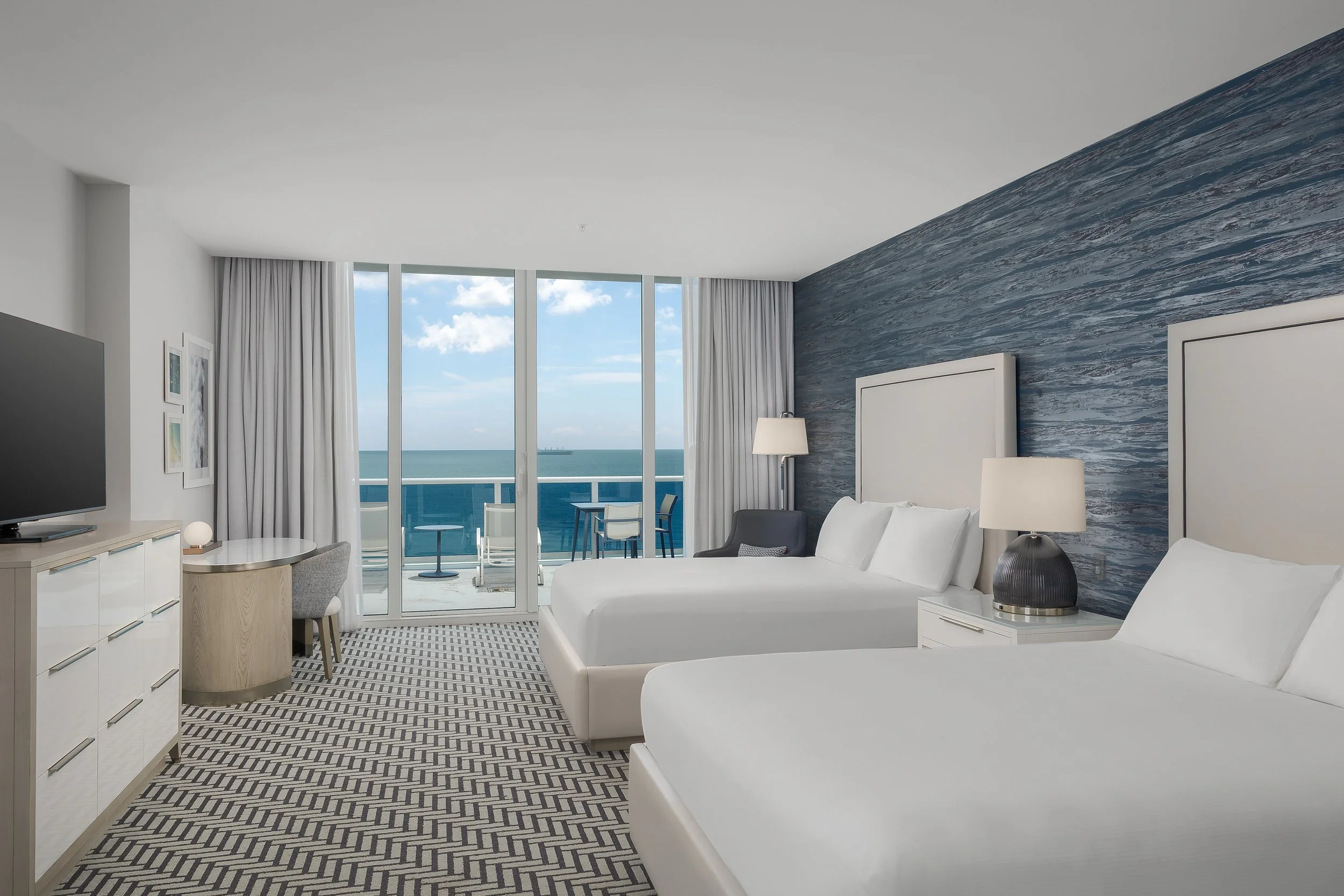 A modern hotel room with white furniture, including two beds, a dresser, and a circular table. It has large sliding glass doors leading to a balcony with outdoor seating, overlooking the ocean with a partly cloudy sky.