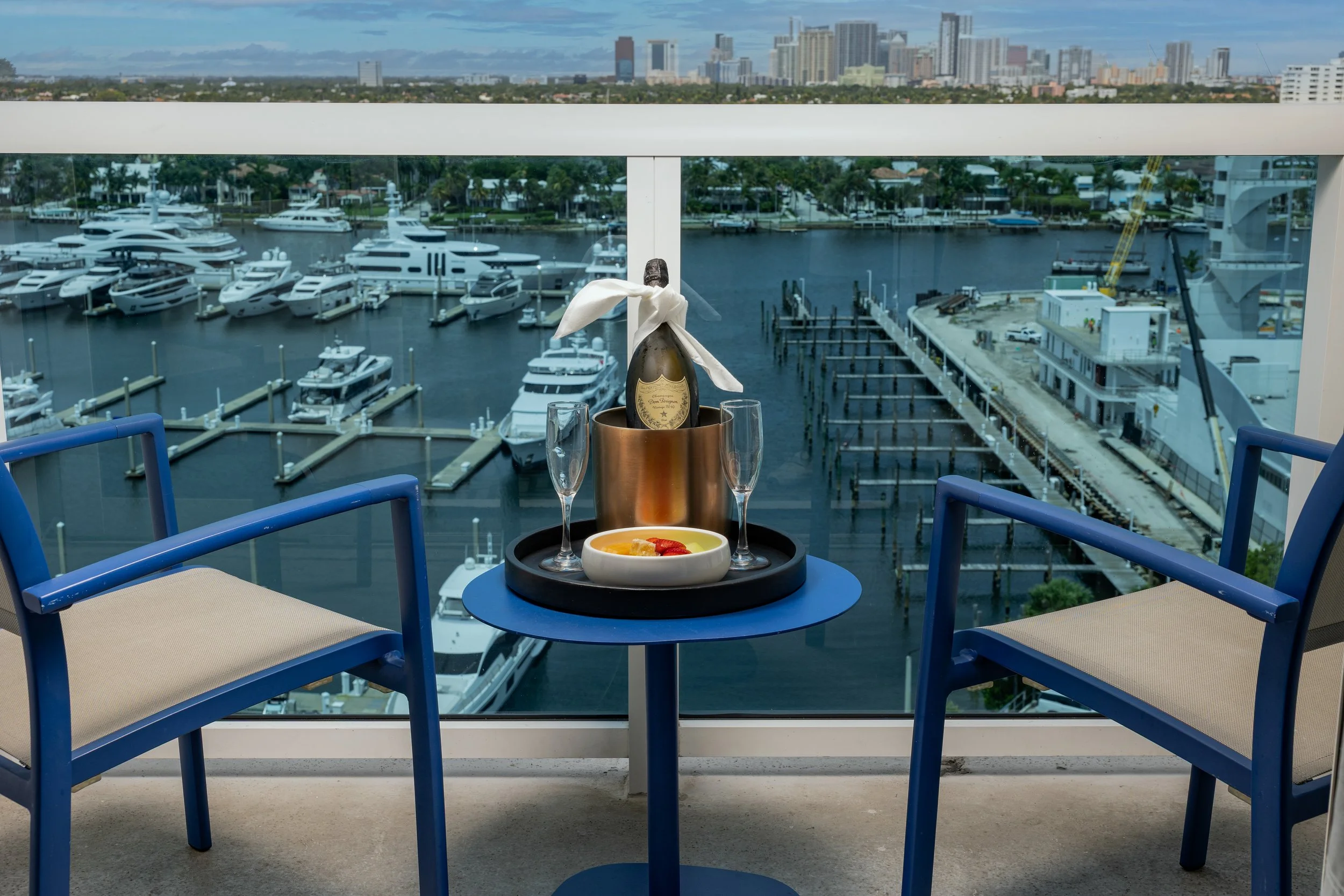 A balcony table set with a bottle of champagne in an ice bucket, two glasses, and a bowl of dessert with strawberries, overlooking a marina with yachts and a city skyline in the background.