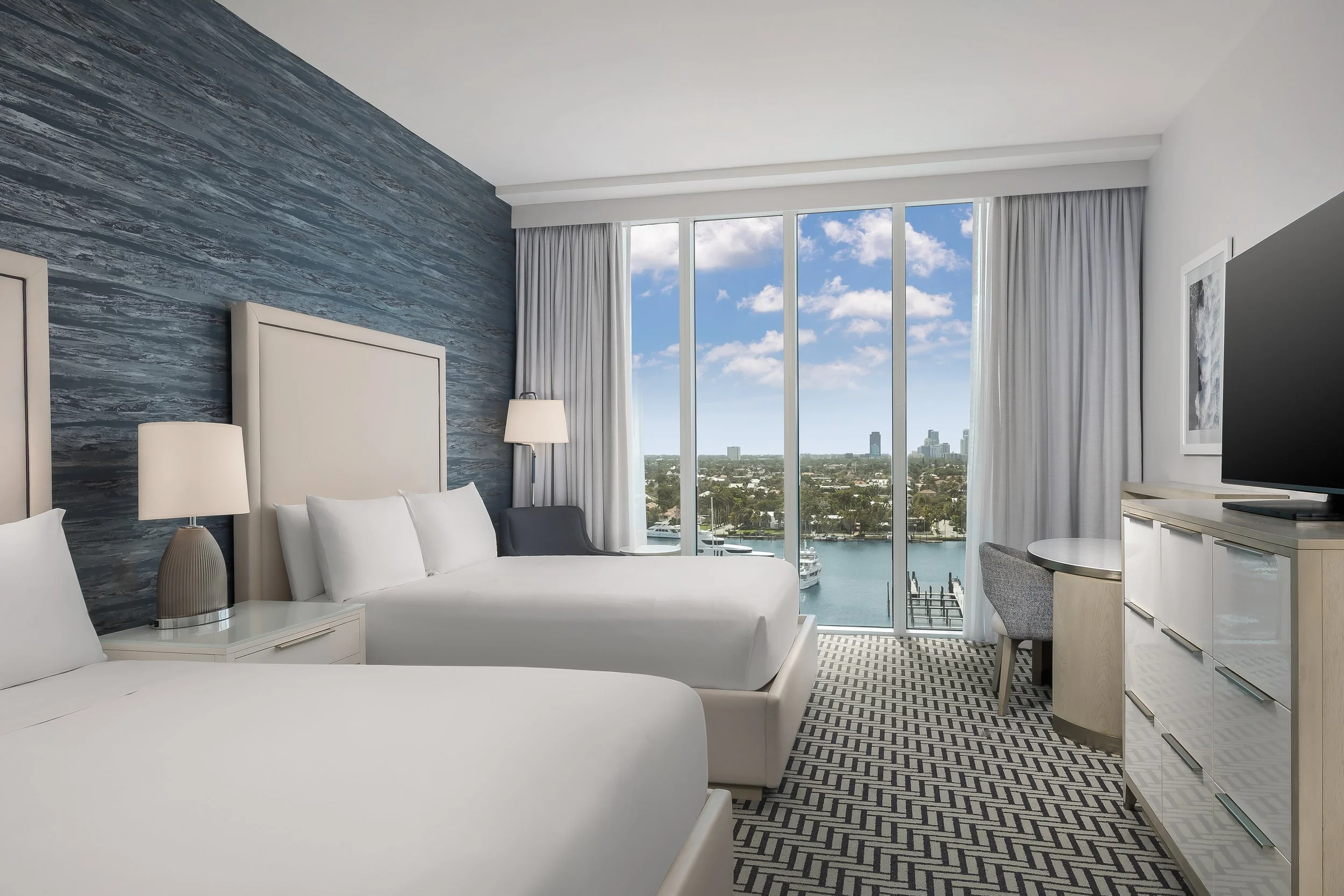 Modern hotel room with white beds, gray patterned carpet, side table with lamp, large floor-to-ceiling window showing city skyline and waterfront, white curtains, TV, and a framed black-and-white photograph on the wall.