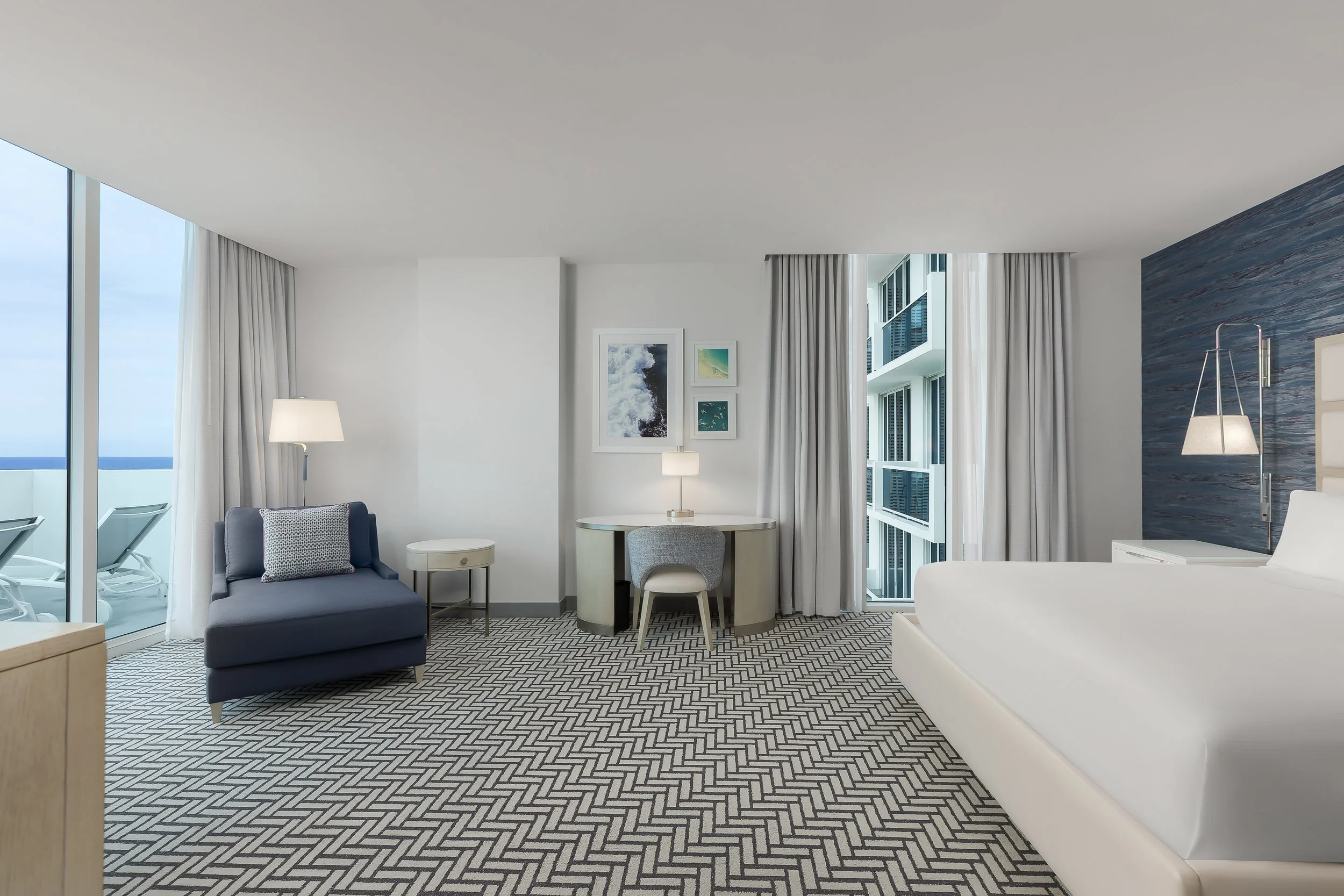 Modern hotel room with white walls, large windows with curtains, a gray armchair with a patterned pillow, a white round table with a small lamp, framed artwork on the wall, and a view of the balcony with ocean.