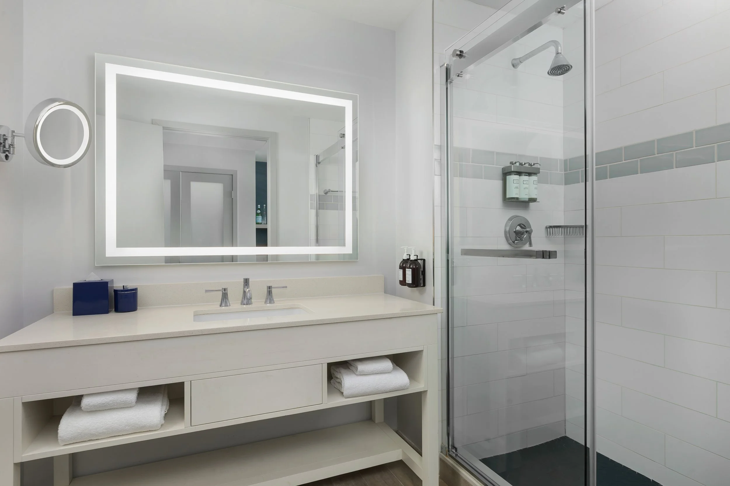 Modern bathroom with a large mirror, vanity with two blue containers, and glass-enclosed shower with shampoo bottles inside.