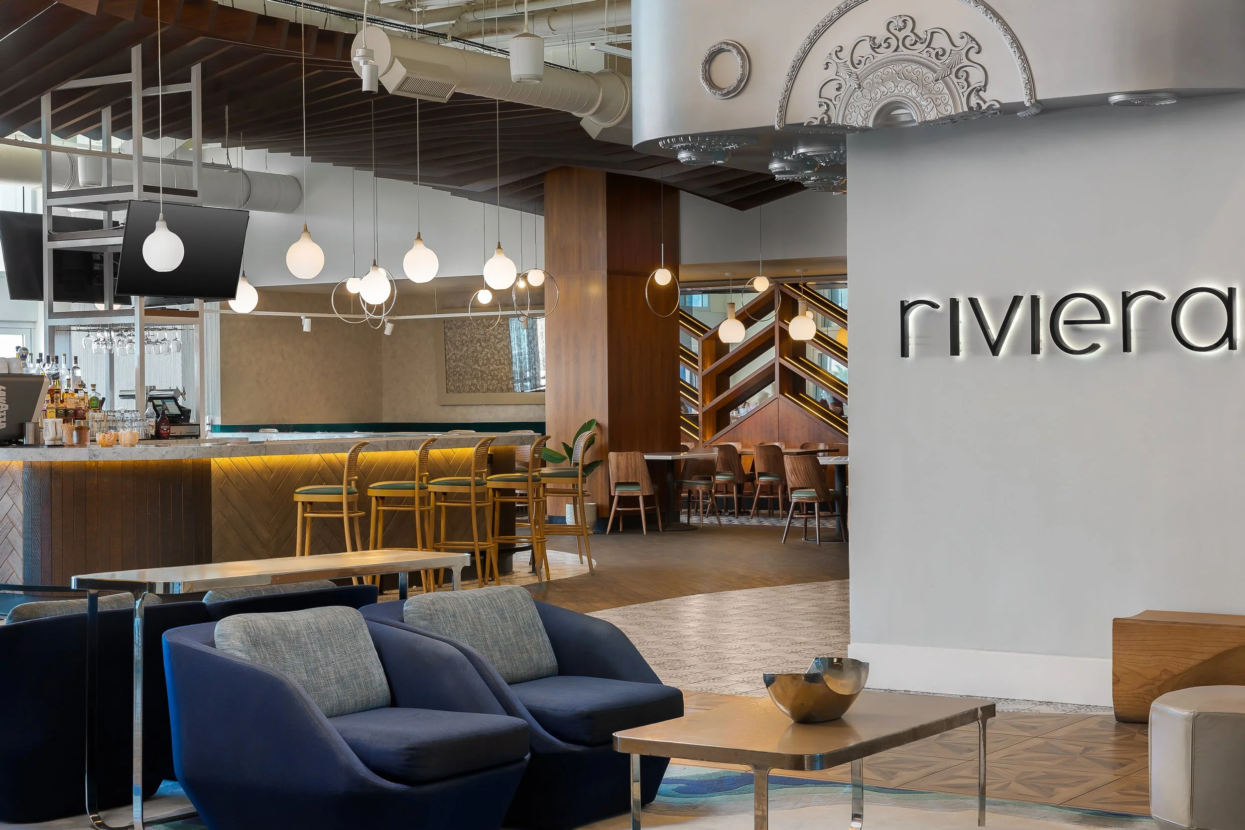 Modern hotel or lounge interior with sitting area, bar, and decorative ceiling elements. Features dark blue chairs, light wood tables, pendant lights, and a white wall with the word 'rivera'.