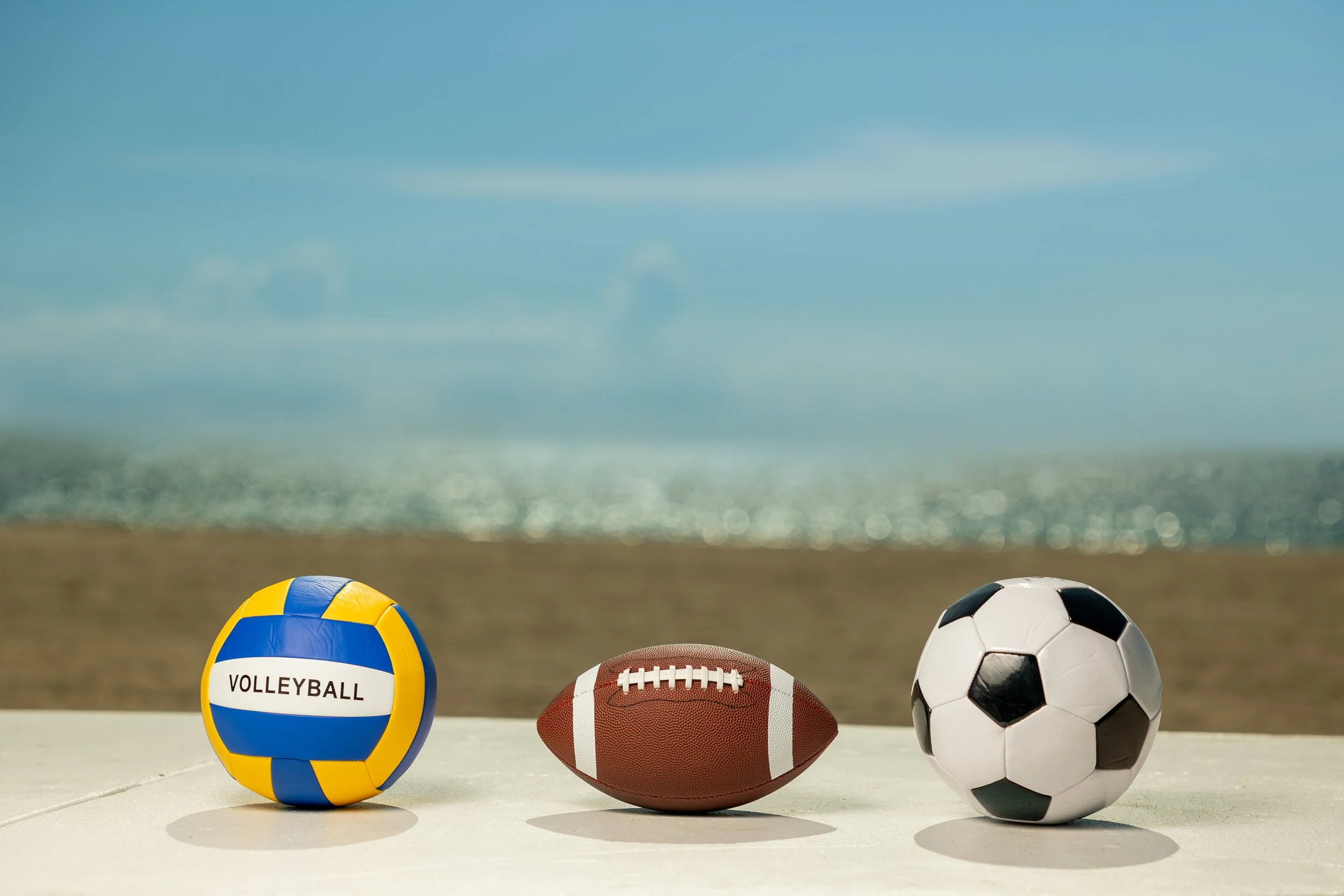 A volleyball, American football, and soccer ball placed side by side on a flat surface outdoors, with a beach and ocean in the background.