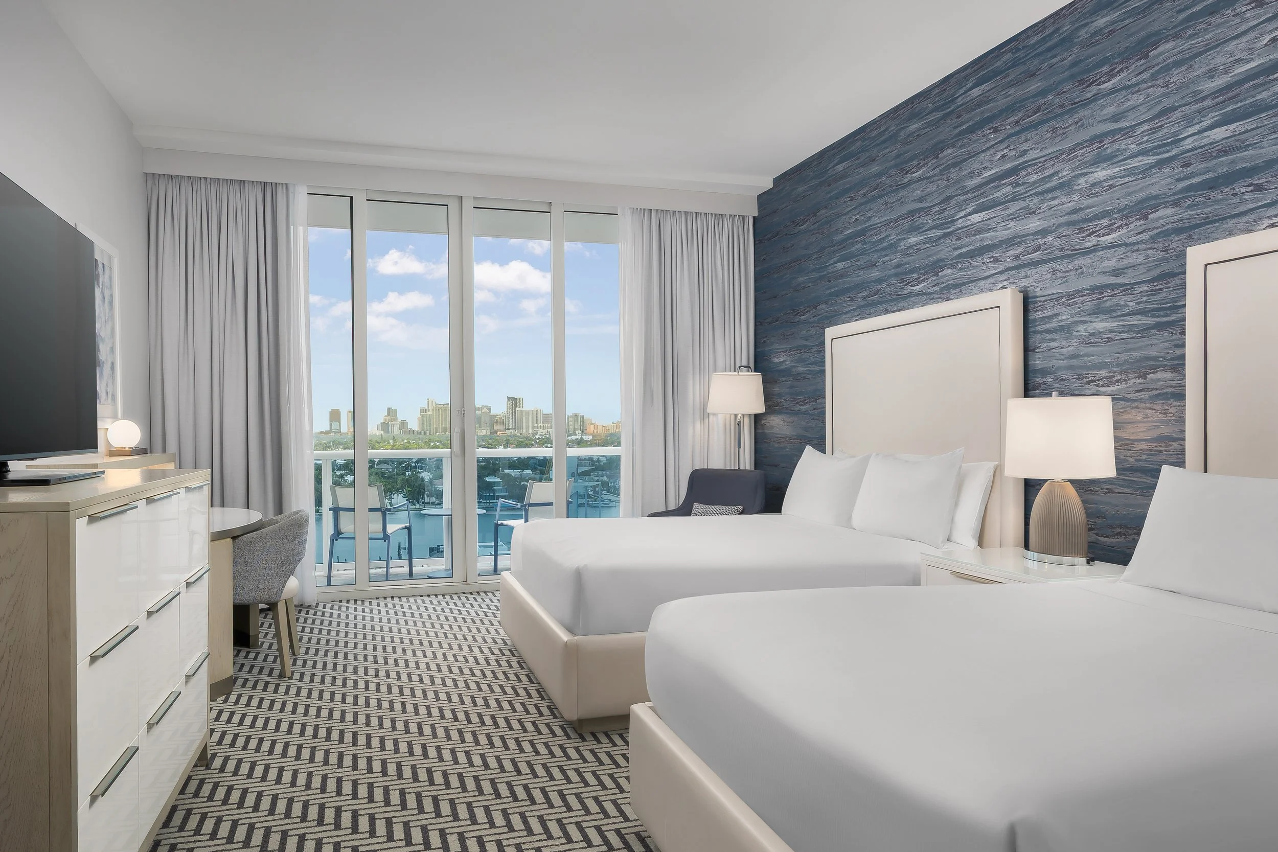 Modern hotel room with large floor-to-ceiling windows showing a city skyline, white beds, a dresser with a lamp, and a balcony with outdoor seating.
