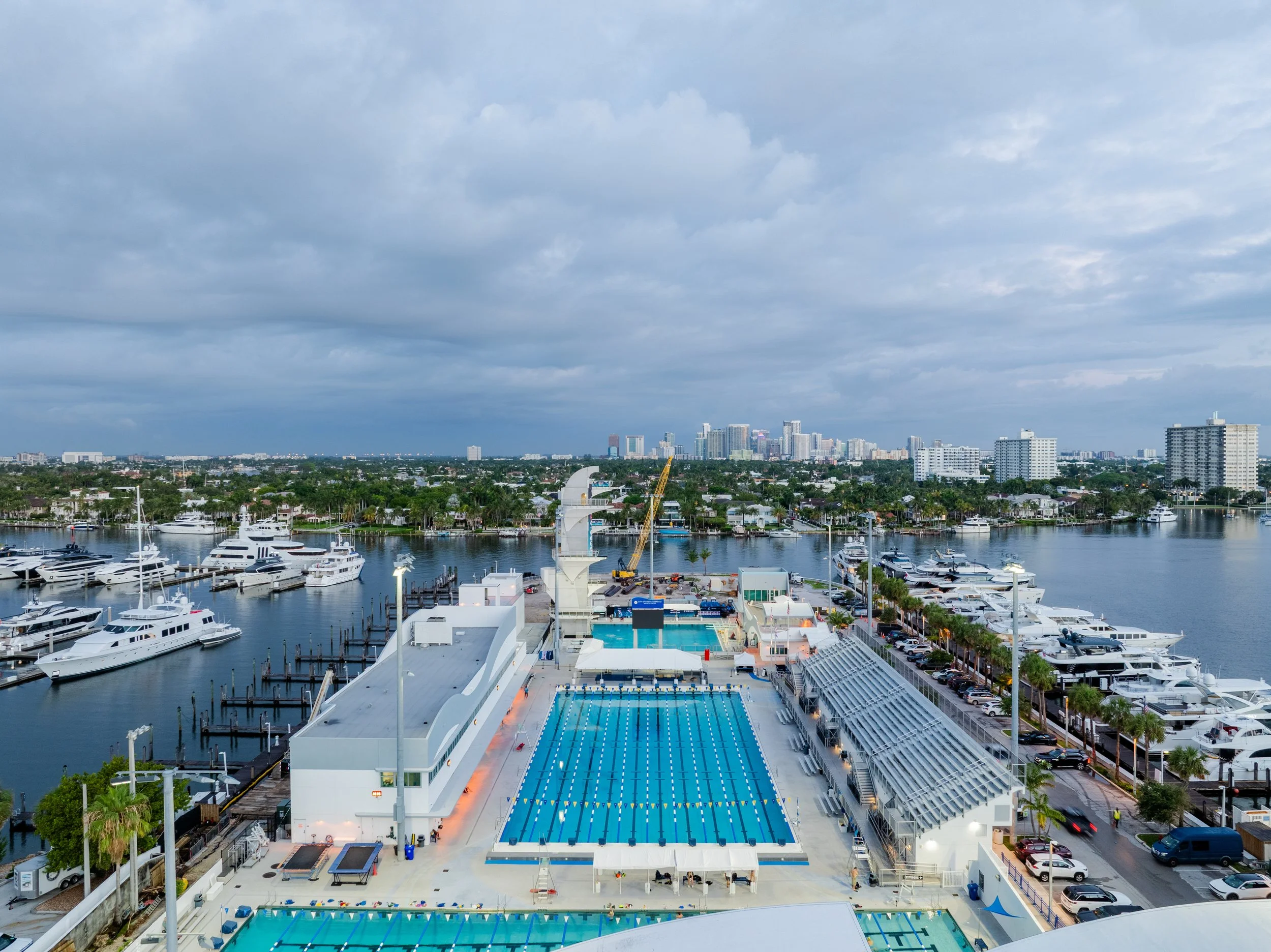 Aerial view of a cityscape with a large outdoor swimming pool, marina with yachts, and high-rise buildings under a cloudy sky.