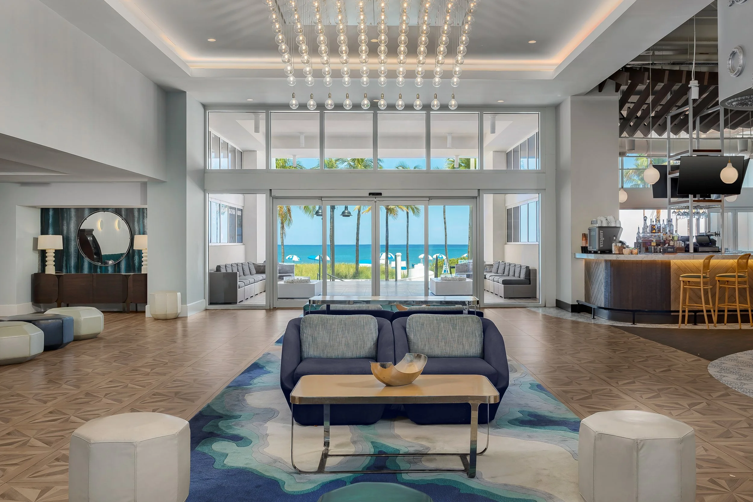 Seaside hotel lobby with large glass doors opening to a beach view with umbrellas, palm trees, and blue ocean. Modern furniture, including sofas, chairs, and a coffee table, decorates the space. Chandeliers and lighting fixtures are suspended from th