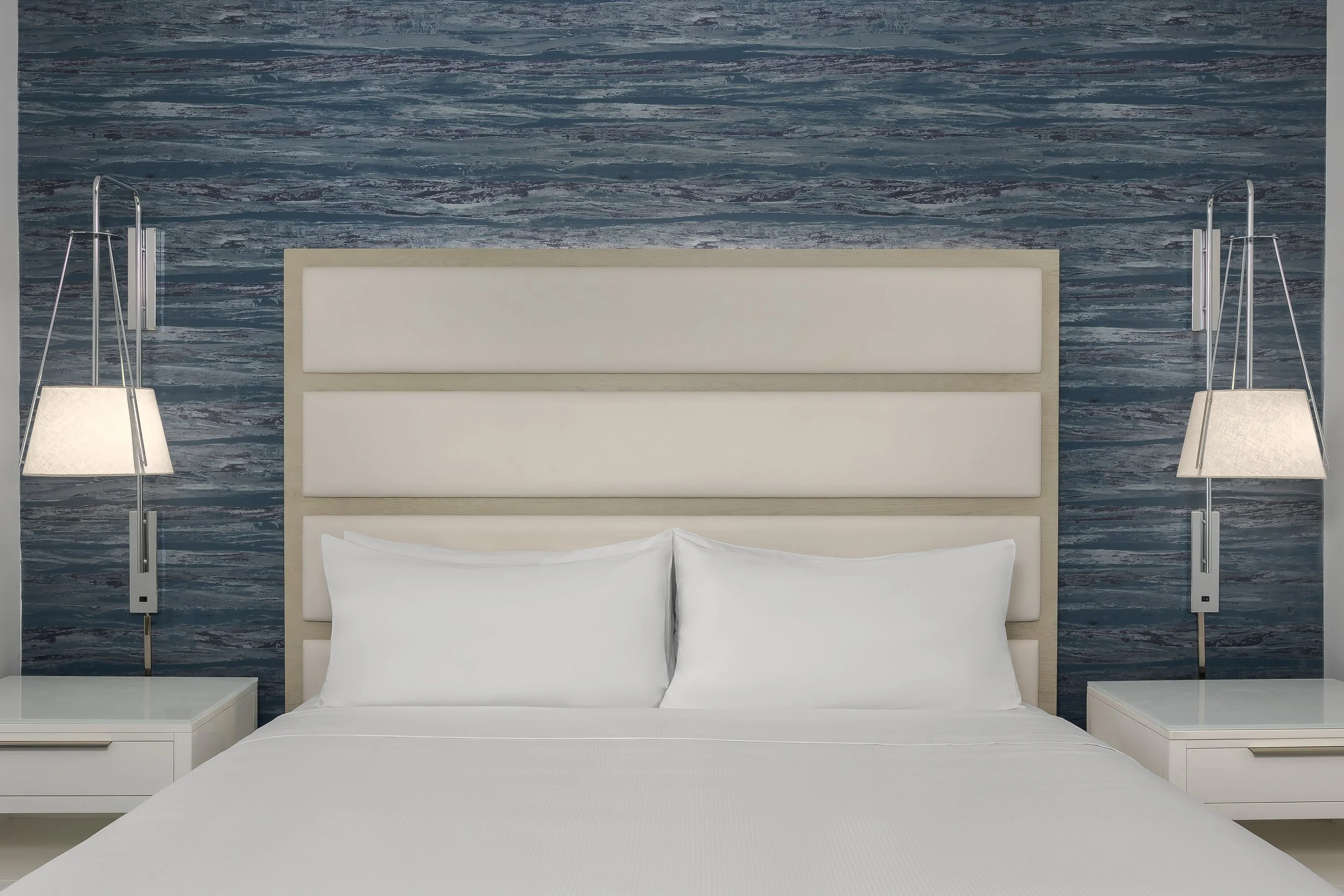 A neatly made bed with white bedding and pillows, flanked by two bedside tables with lamps. The headboard is beige and padded, set against a textured blue wall.