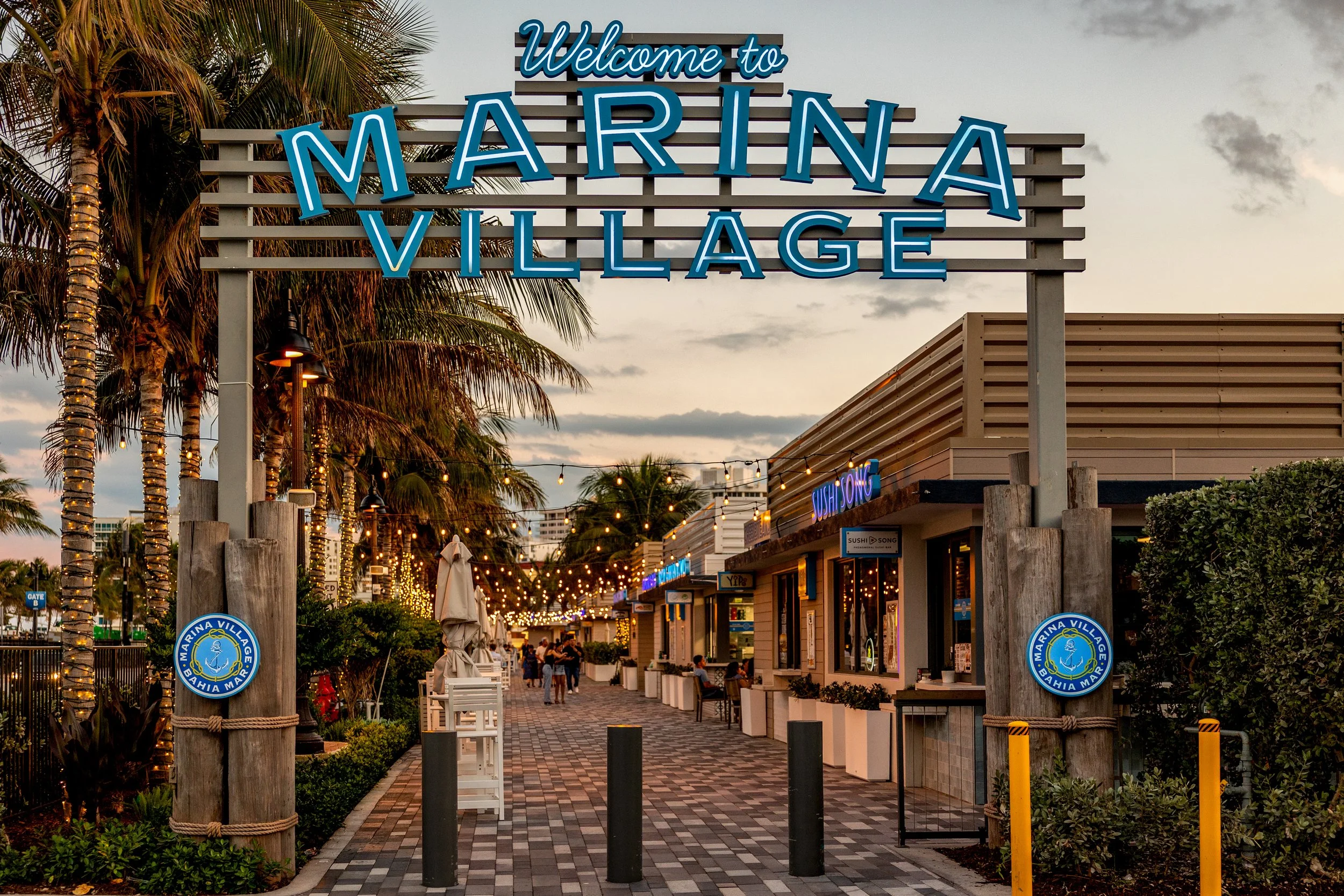 The image shows a welcoming sign for Marina Village in Bahia Mar, outdoors with palm trees and string lights. There are shops and people walking in the background during evening or dusk.