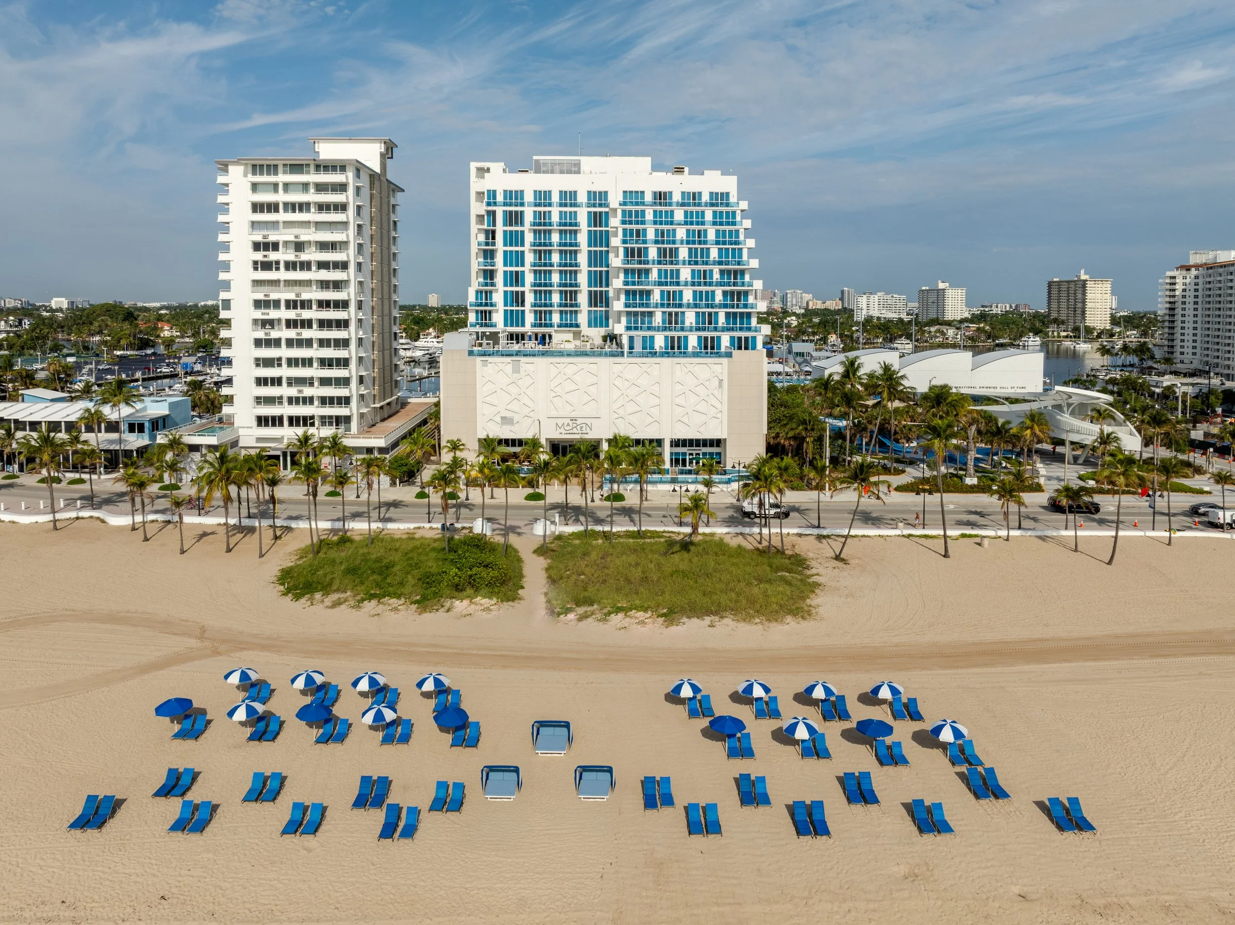 Beach with blue umbrellas and lounge chairs, sandy shore in foreground, tall modern buildings and marina in background under partly cloudy sky.