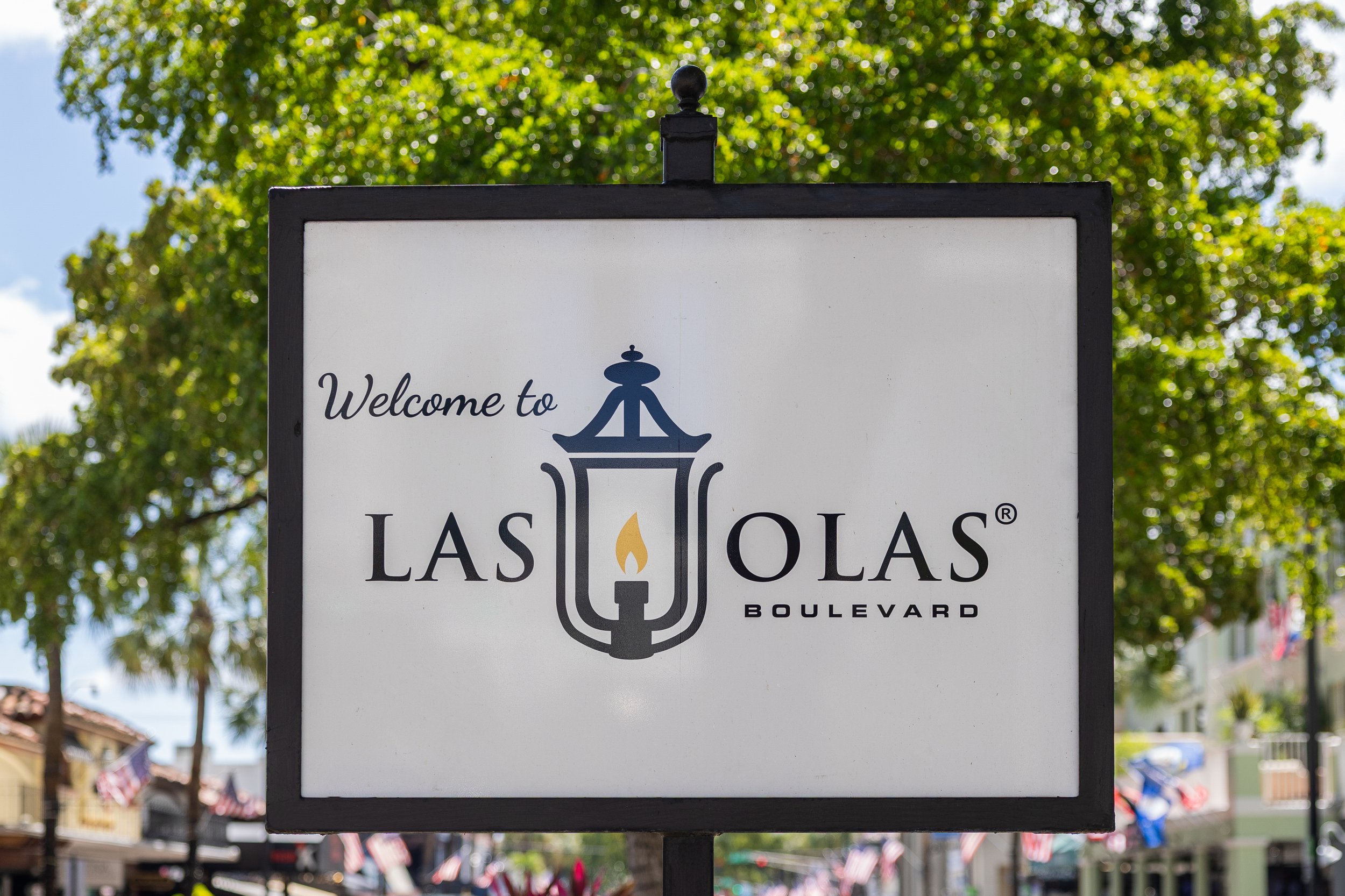A welcome sign for the Las Olas Boulevard in Fort Lauderdale, Florida. The sign includes text and a graphic of a streetlamp with a flame, set against a backdrop of green trees and clear blue sky.
