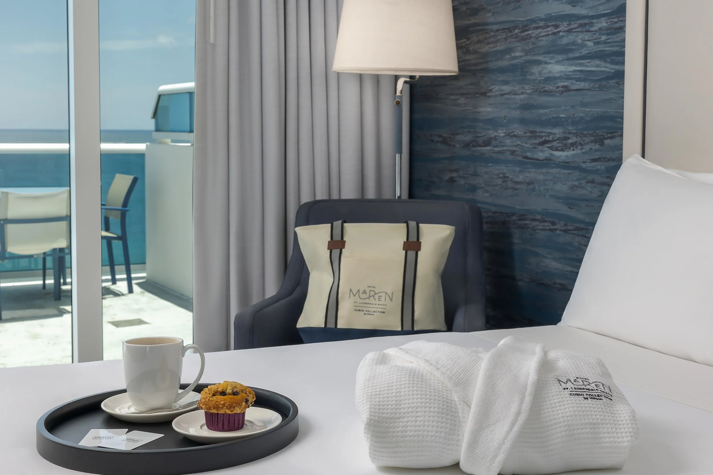A hotel room with a bed, a tray with a muffin and a cup of coffee, a reusable tote bag, and white towels with hotel branding, overlooking a balcony with outdoor chairs and an ocean view.