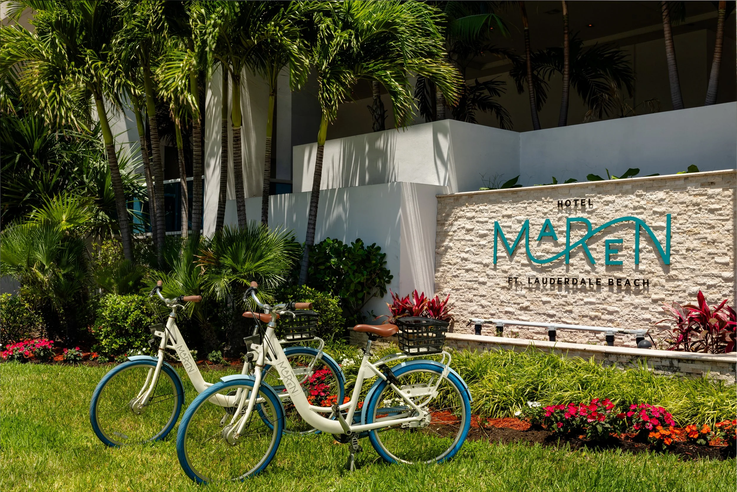 Bicycles parked on a grassy area near a garden with pink flowers, in front of a hotel sign that reads 'Hotel Maken Ft. Lauderdale Beach' surrounded by lush green palm trees.