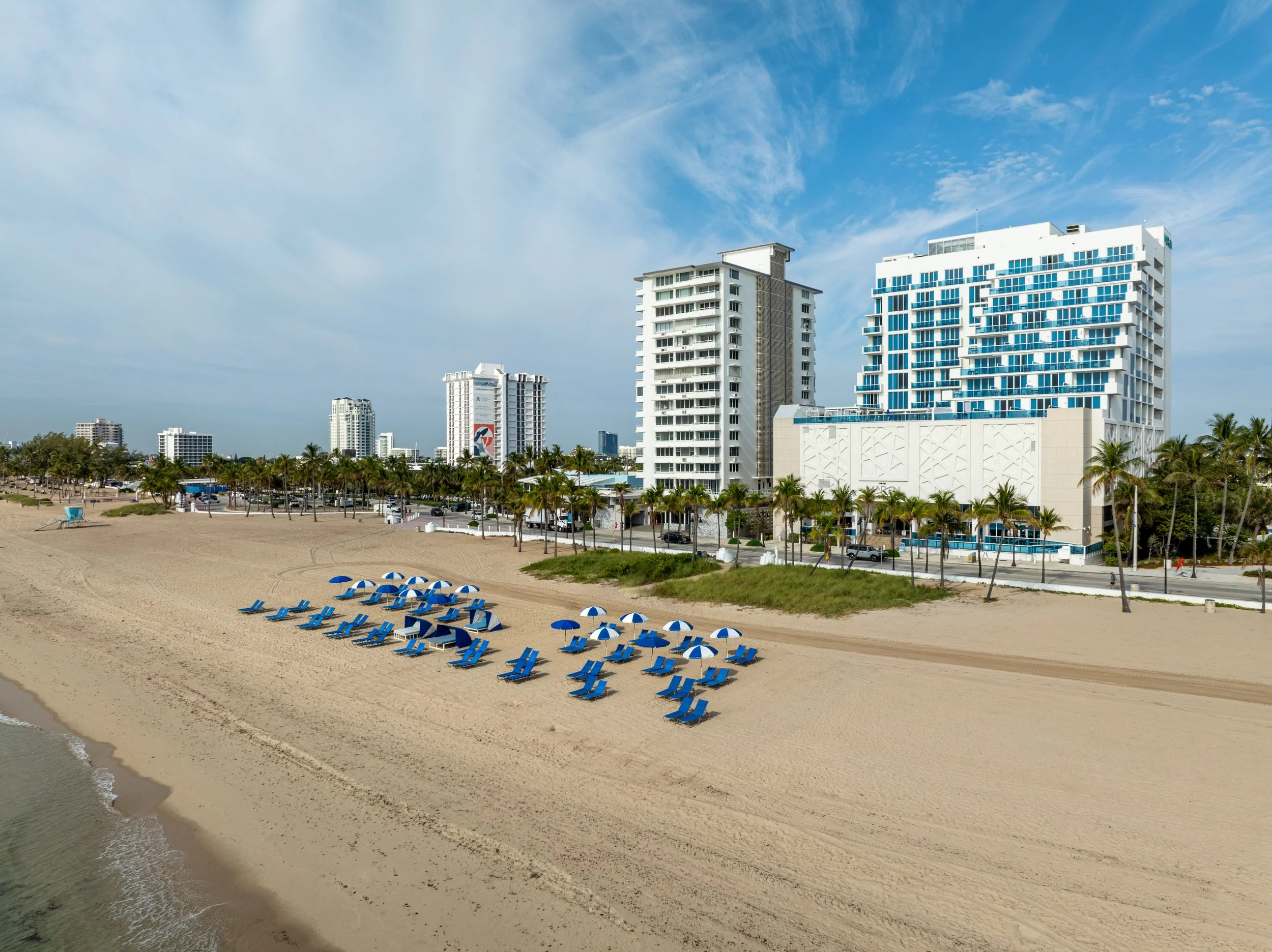 Empty blue beach umbrellas and lounge chairs on a sandy beach with high-rise buildings and palm trees in the background.