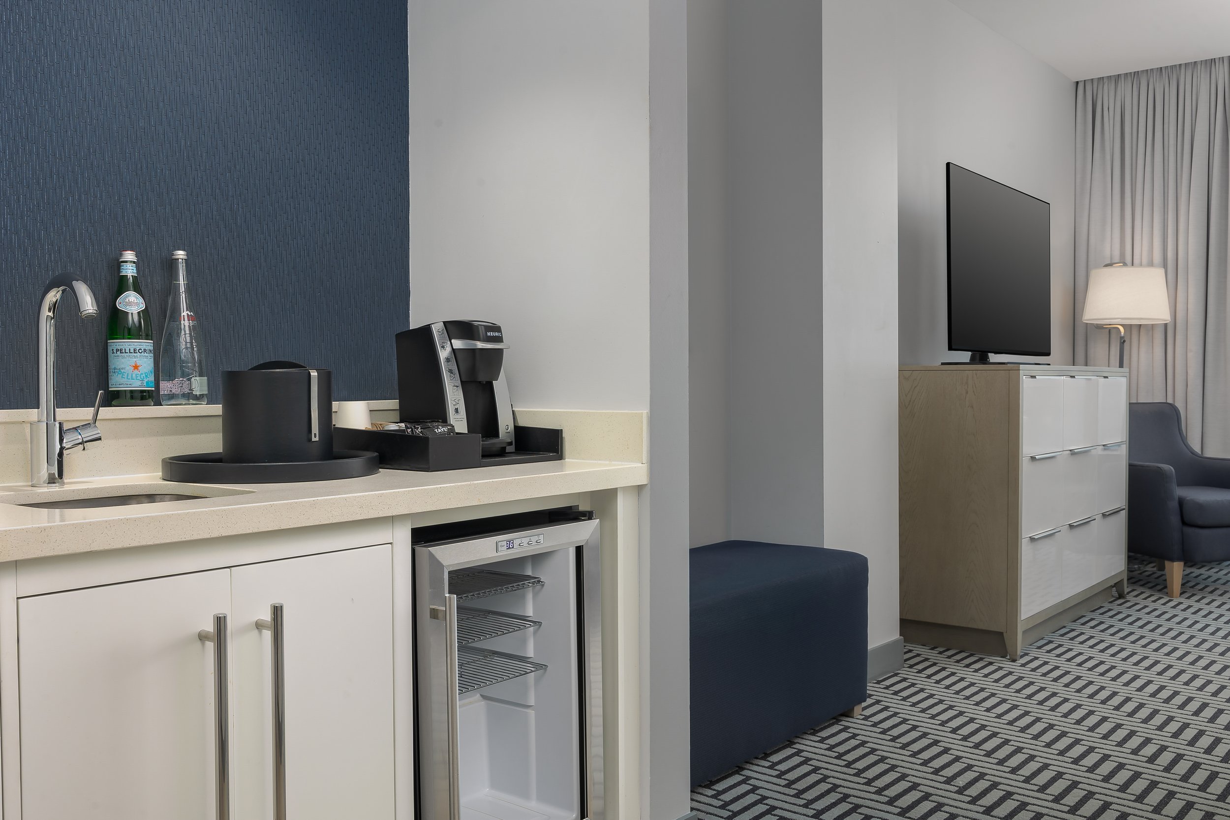A kitchenette area with a sink, a hot water kettle, a coffee maker, and bottled water on a counter. In the background, there is a TV, a lamp, and a chair in a hotel room.