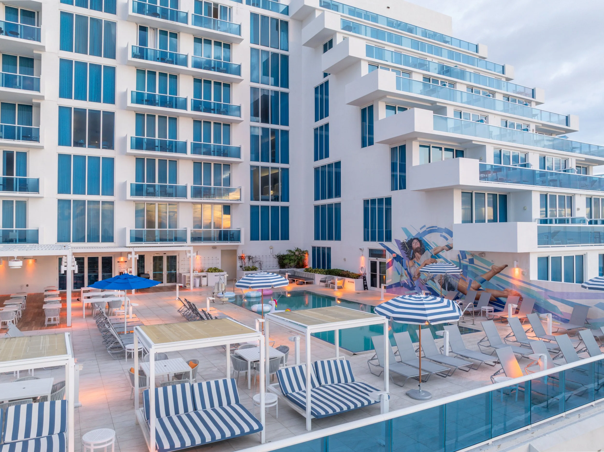 Exterior view of a modern hotel with a swimming pool, lounge chairs with blue and white striped cushions, and blue and white umbrellas. In the background, there is a tall white building with large glass windows and balconies, and a cloudy sky.