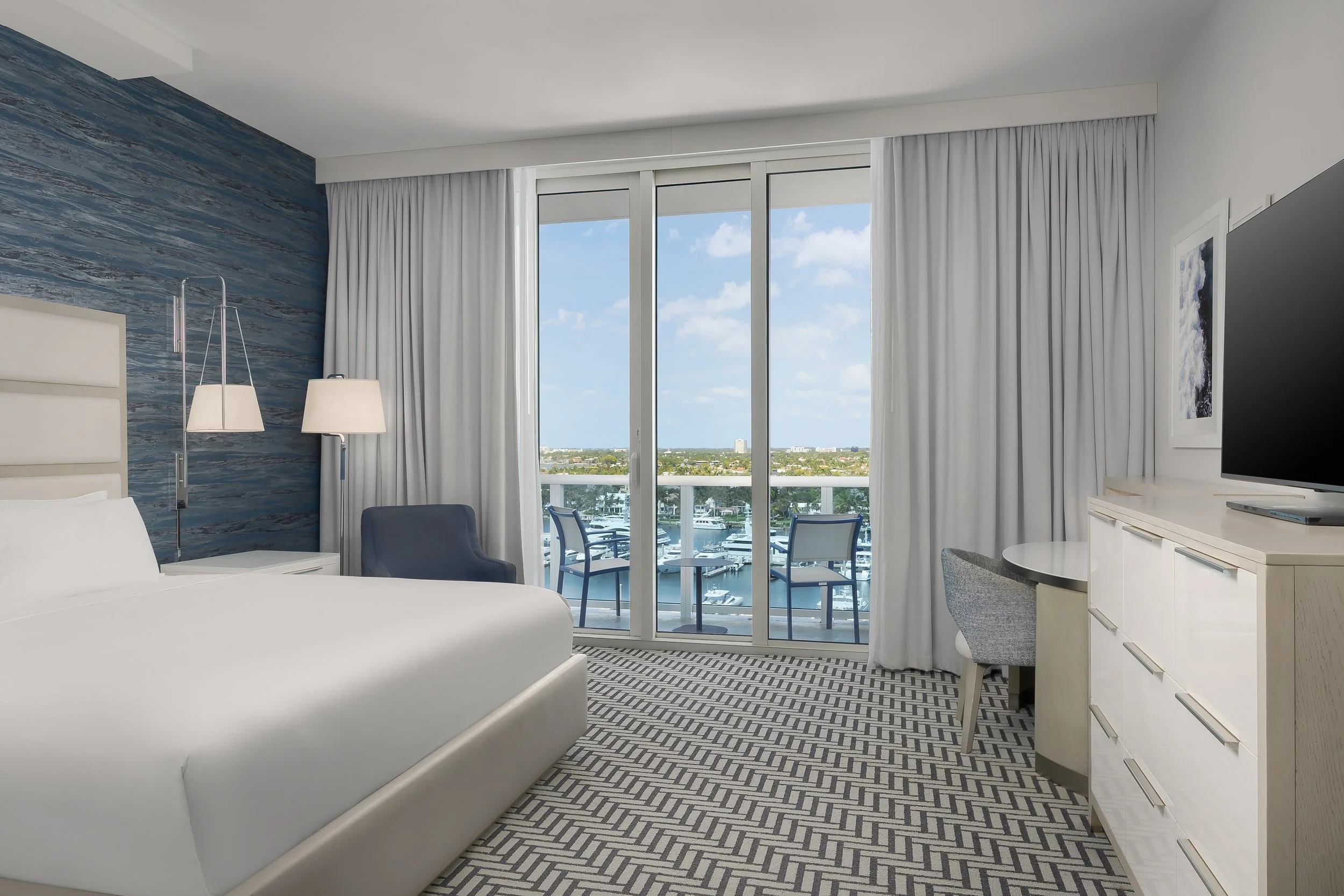 Interior of a modern hotel room with a white bed, blue accent wall, floor lamps, sliding glass doors leading to a balcony with outdoor seating, city and water view, white curtains, and a TV on a white dresser.