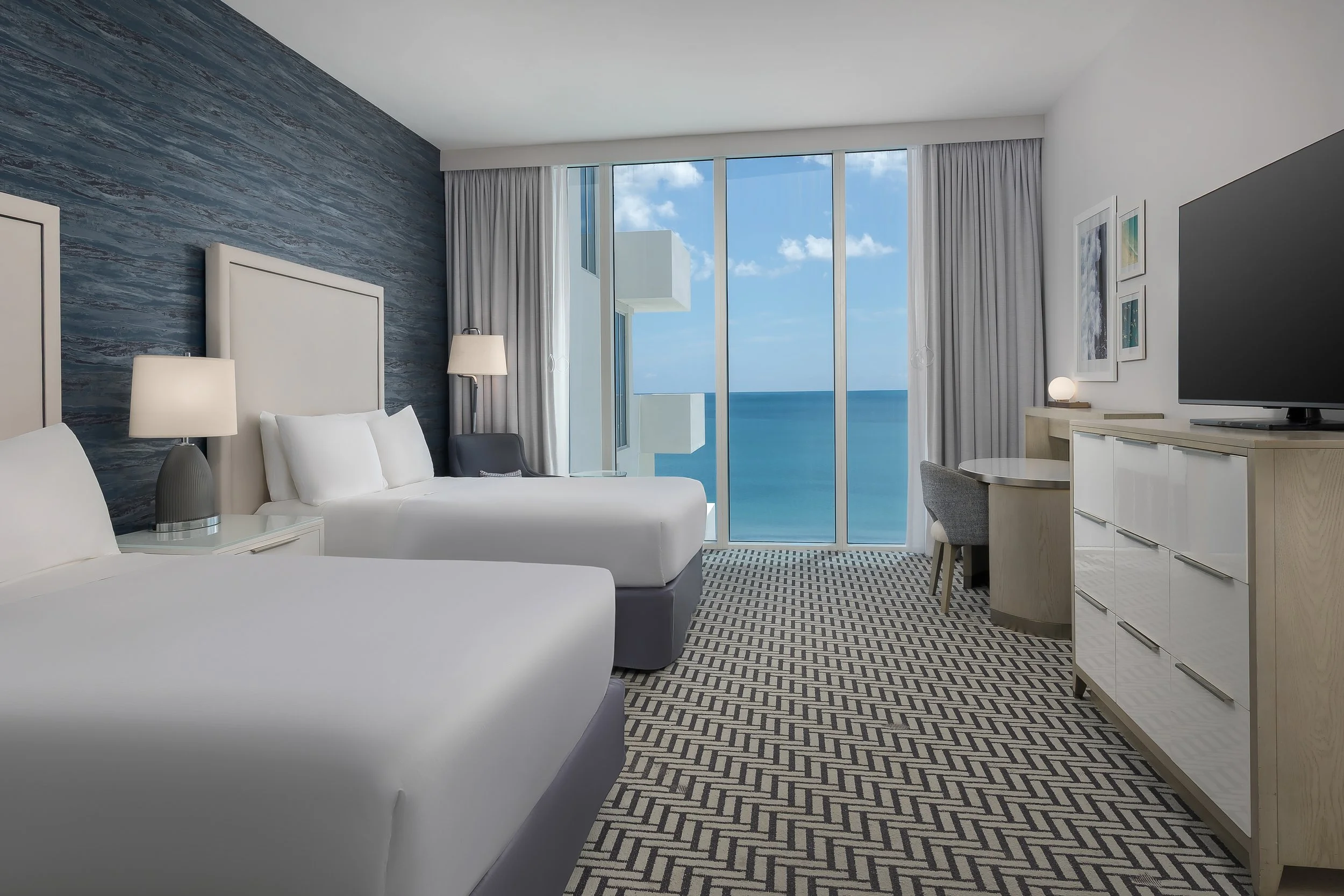 Modern hotel room with coastal view, twin beds, white headboards, bedside lamps, large window with white curtains, ocean view, wall art, television, dresser, and a patterned carpet.