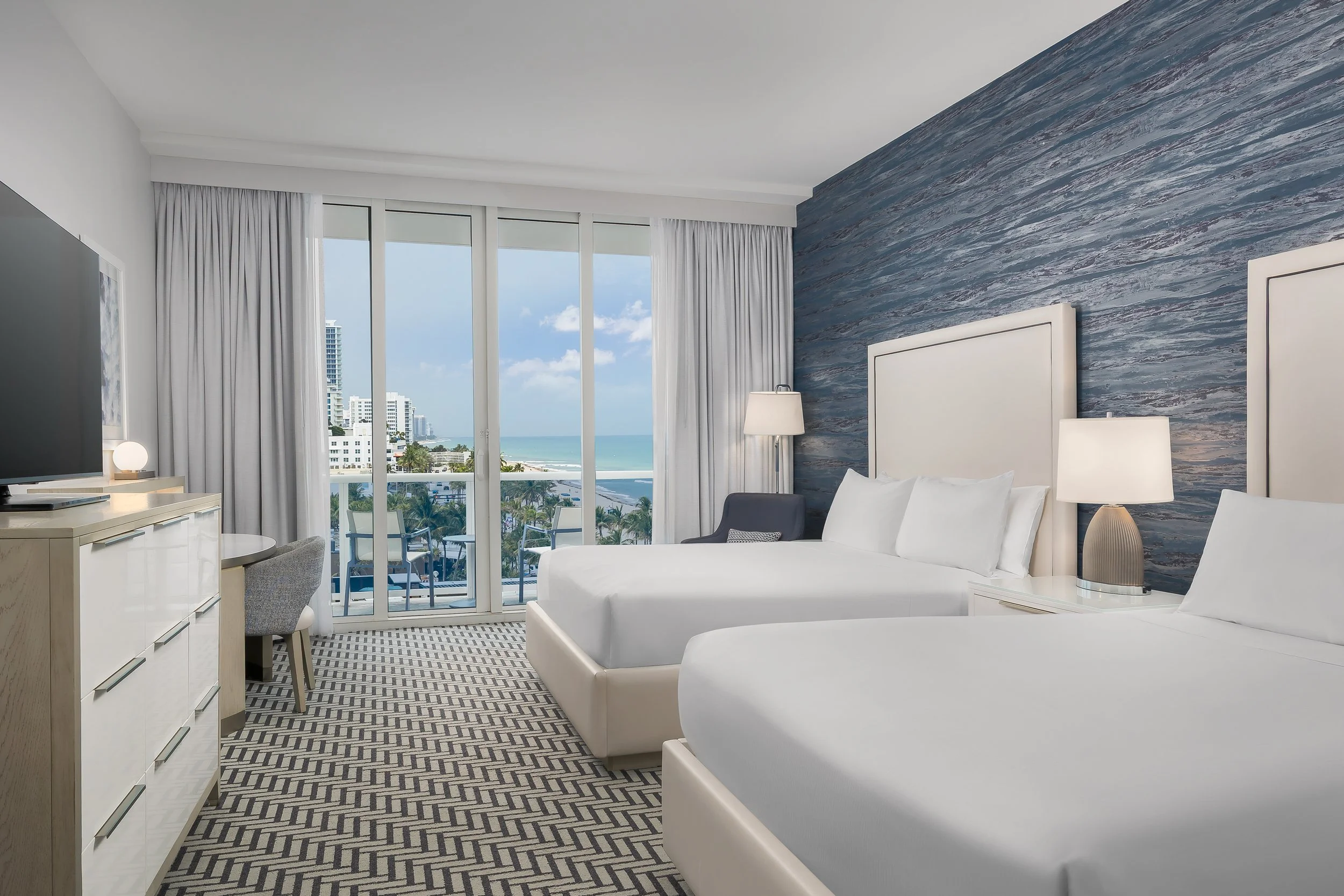 Hotel room with a view of the ocean, featuring white furniture, a flat-screen TV, a lamp, a patterned carpet, large windows with white curtains, and a balcony with outdoor seating.