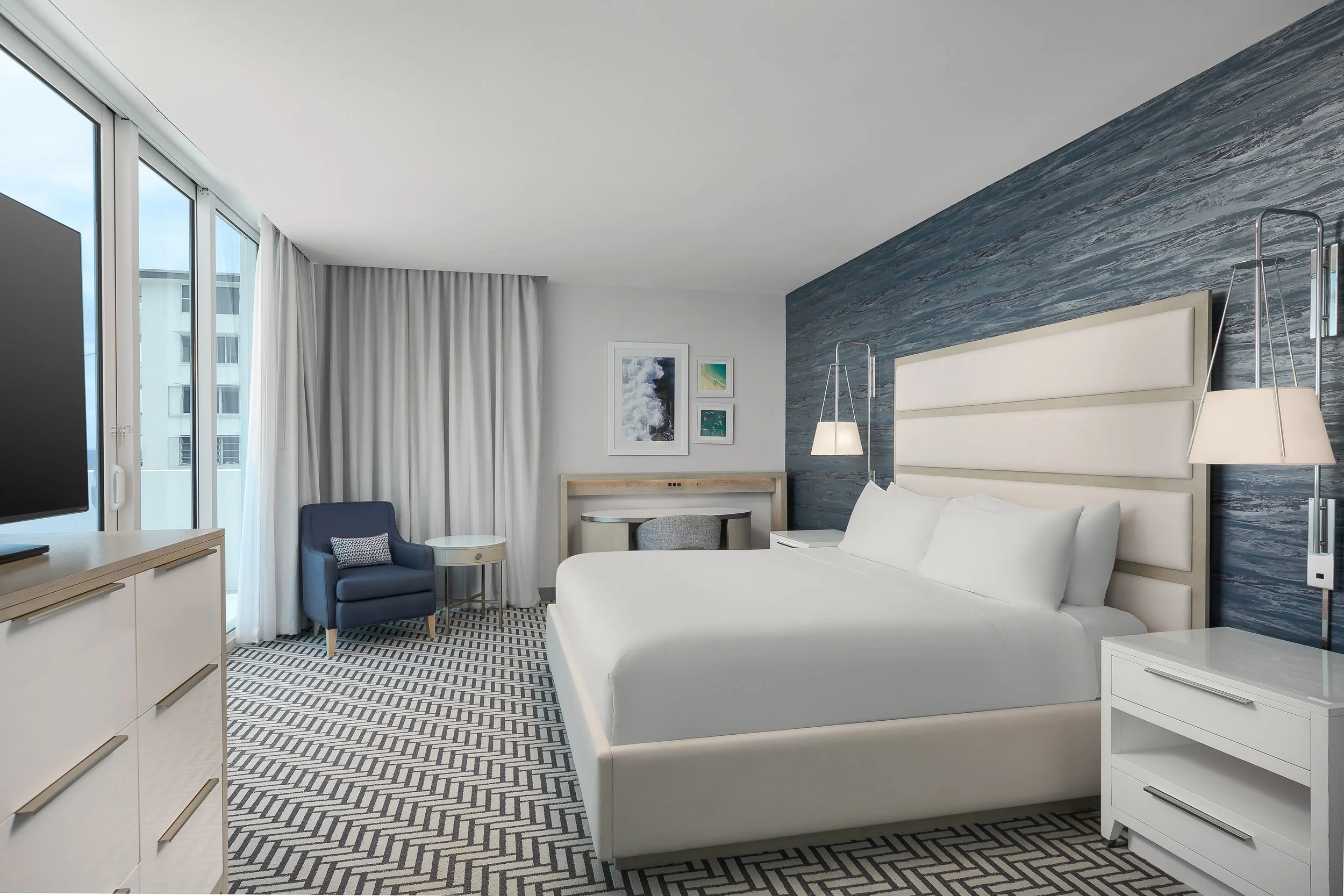 Modern hotel room with a large bed, ocean-themed wall art, armchair, side tables, and floor-to-ceiling windows with curtains